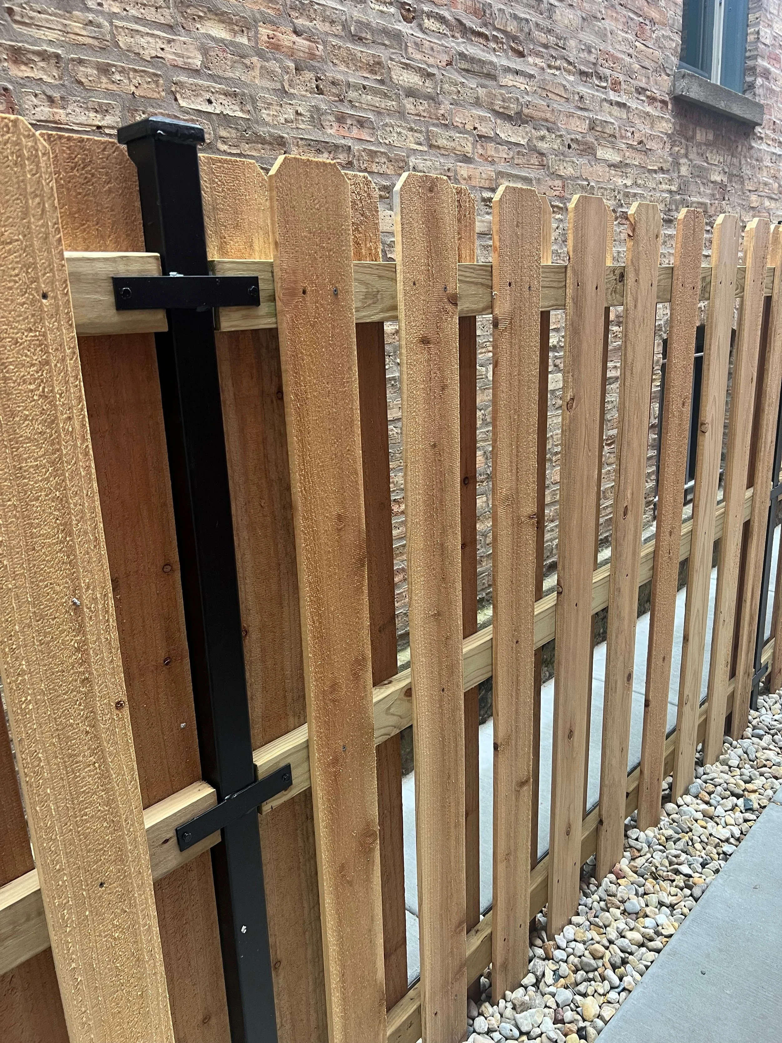 A black gate latch attached to a light brown wooden fence in front of a brick building wall, with a window visible in the top right corner and a small section of a concrete sidewalk and pebbled ground on the right side.