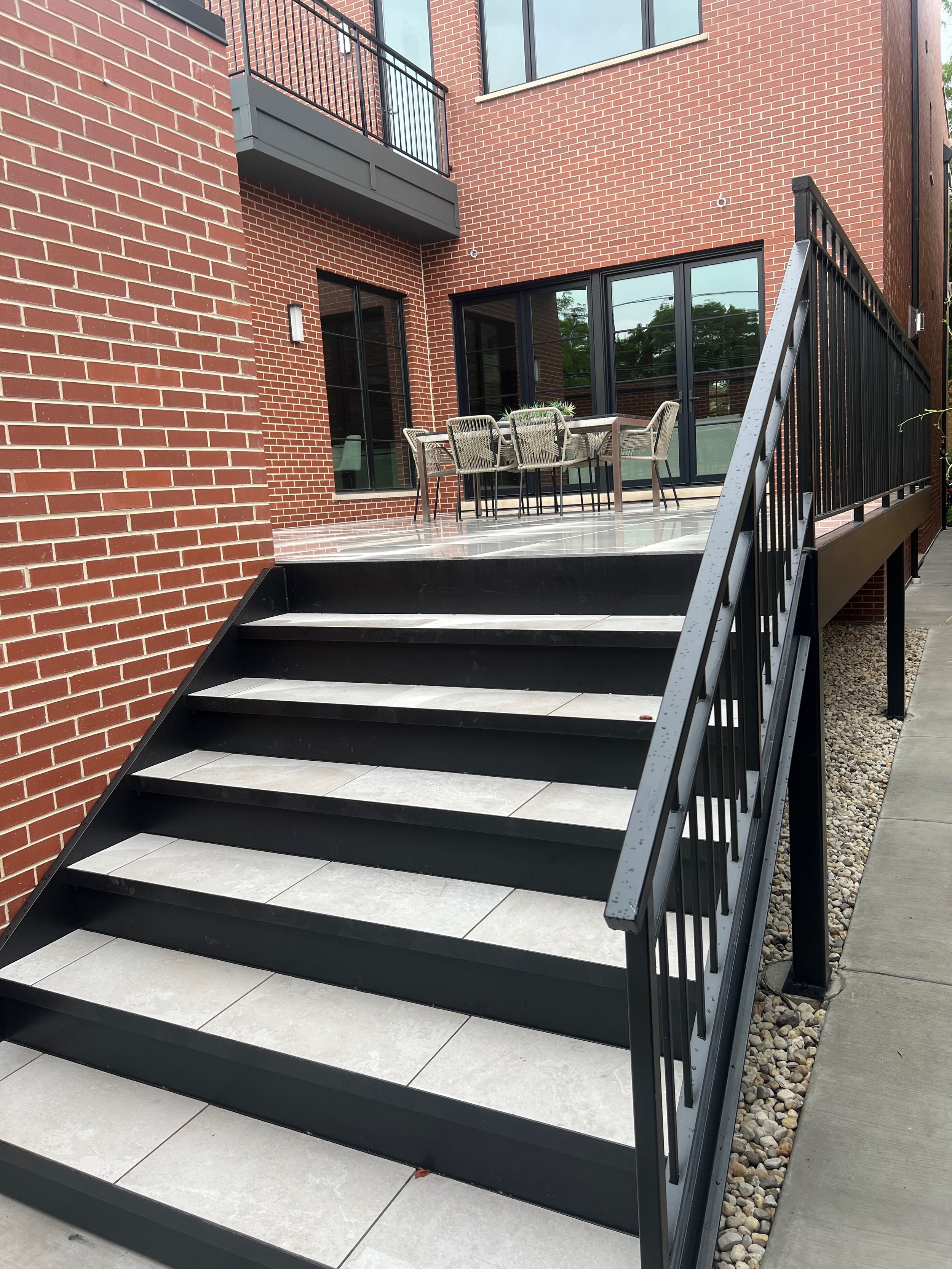 A modern apartment building with a black metal staircase leading up to a balcony and patio area with a table and chairs. The building has red brick walls and large glass windows.