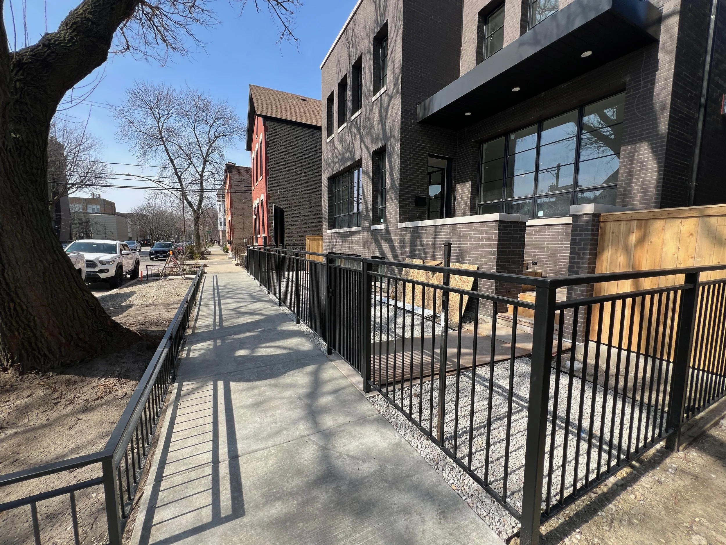 A sidewalk runs alongside modern black brick residential buildings, with a wooden fence and black metal railing. Cars are parked along the street, and leafless trees cast shadows on the buildings under a clear blue sky.