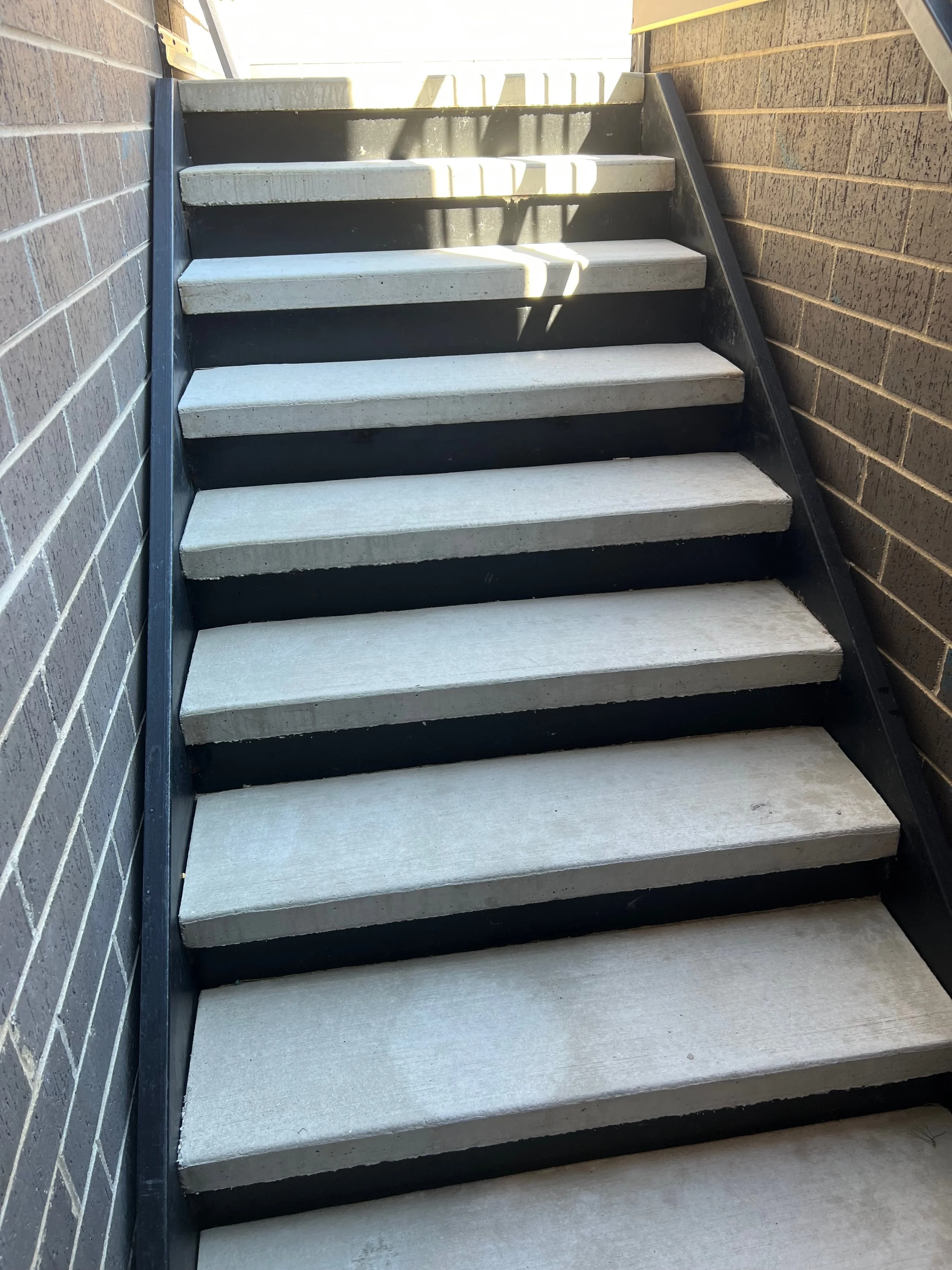Concrete stairs with black sides, situated between brick walls on either side.