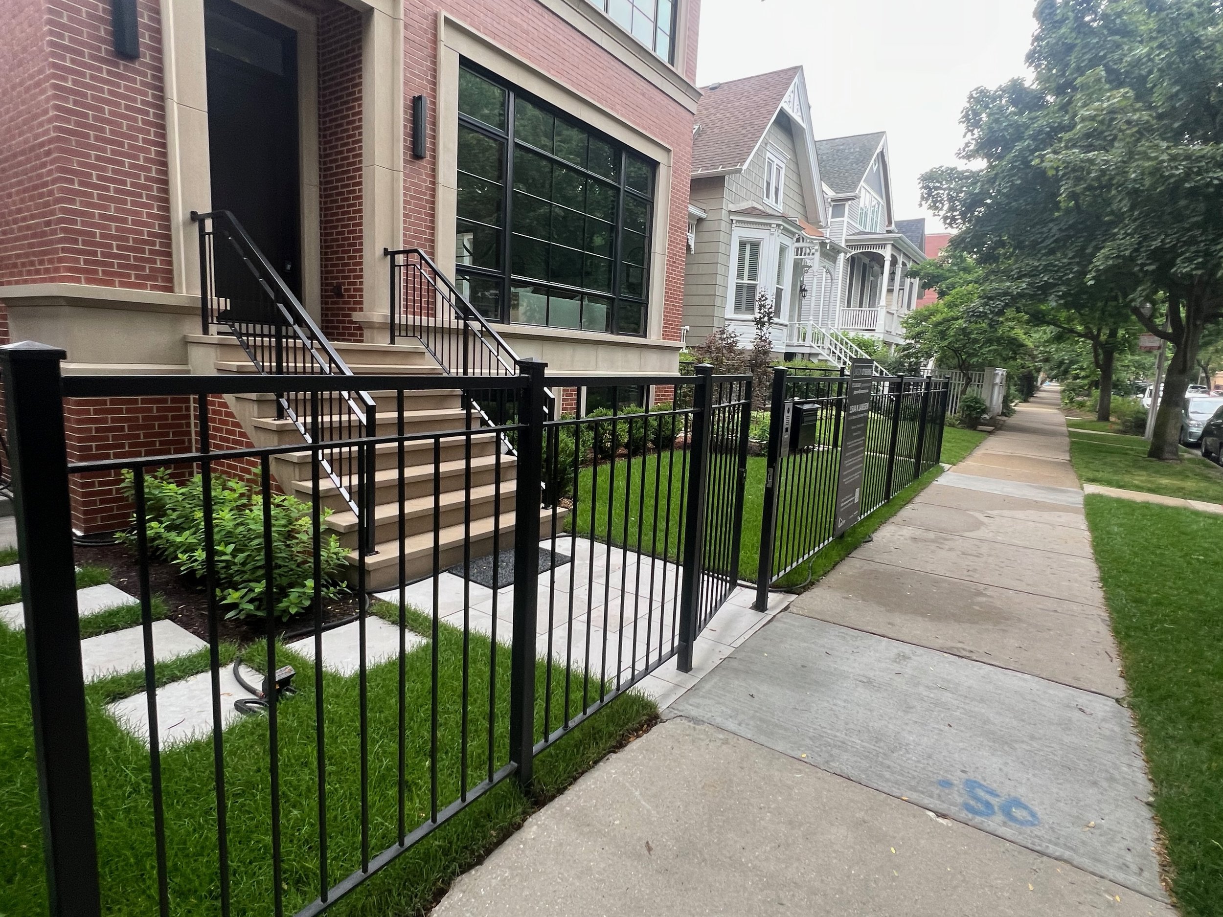 Residential street with hybrid townhouses featuring brick and siding exteriors, front stairs, black metal fences, trees, sidewalk, and parked cars.