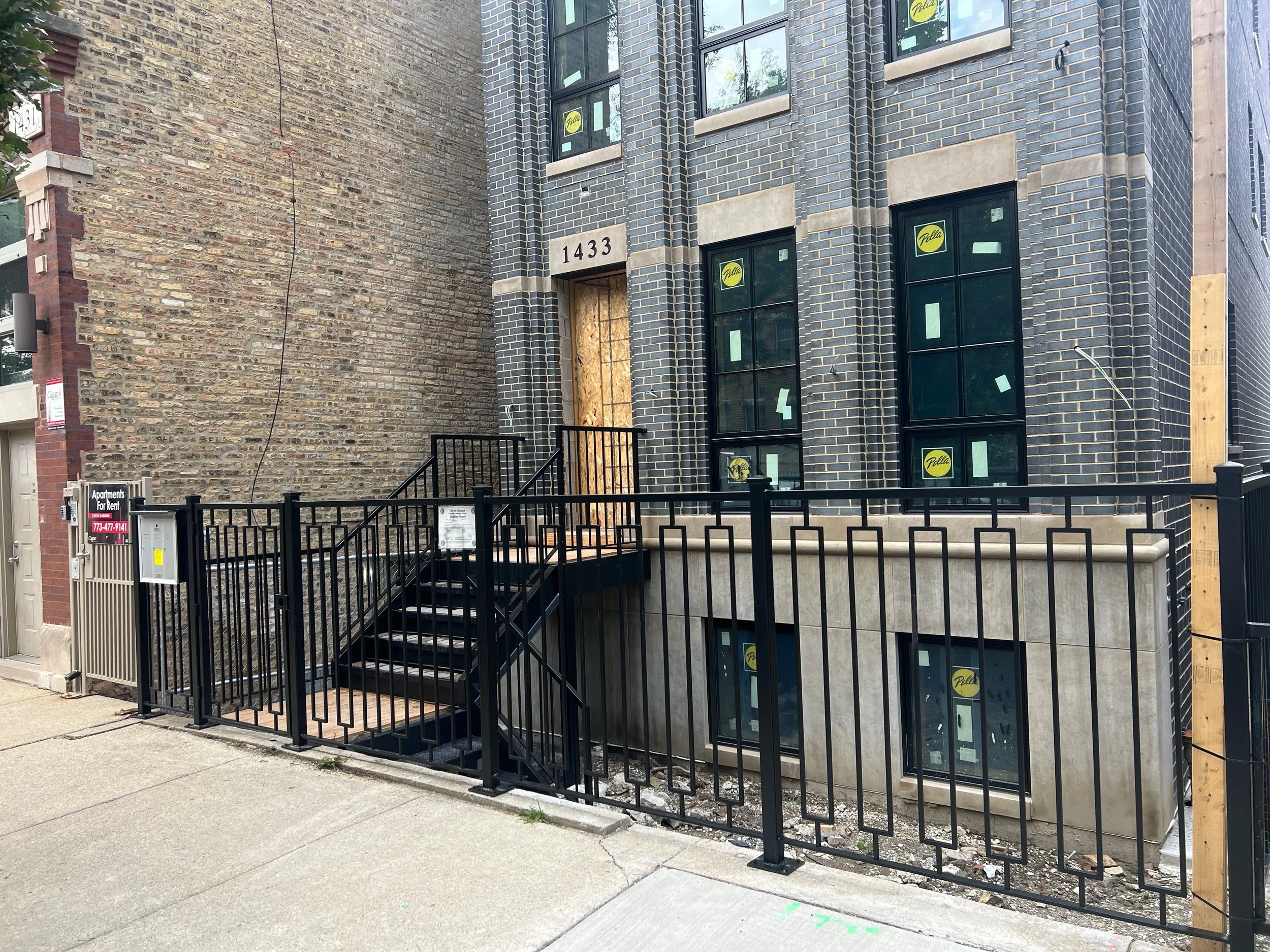 Construction site with a building under renovation, exposed brick and unfinished windows, black metal gate, and sidewalk in the foreground.