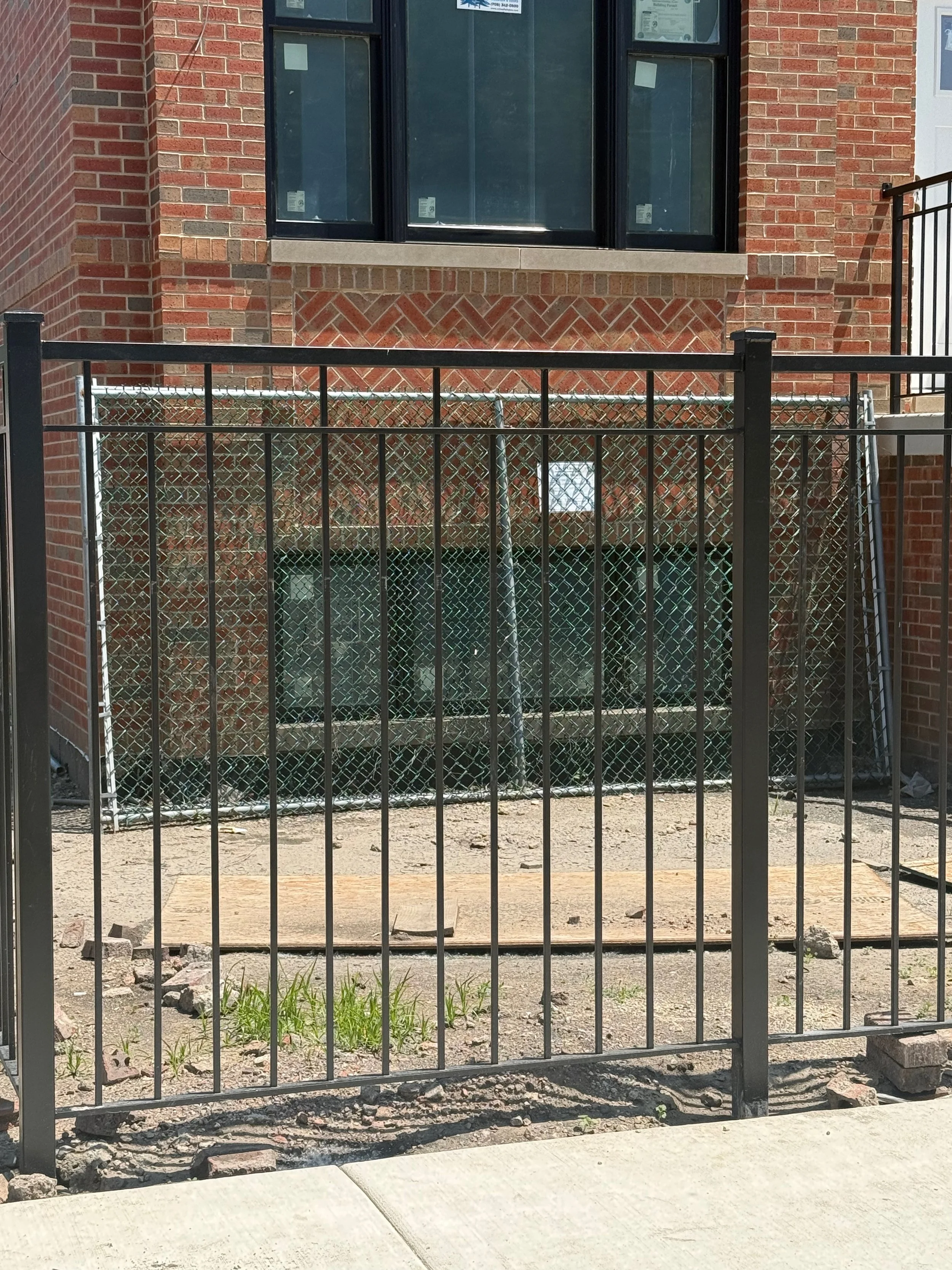 A black metal fence in front of a construction site with a brick building in the background, a window, and an open area with dirt and small plants.