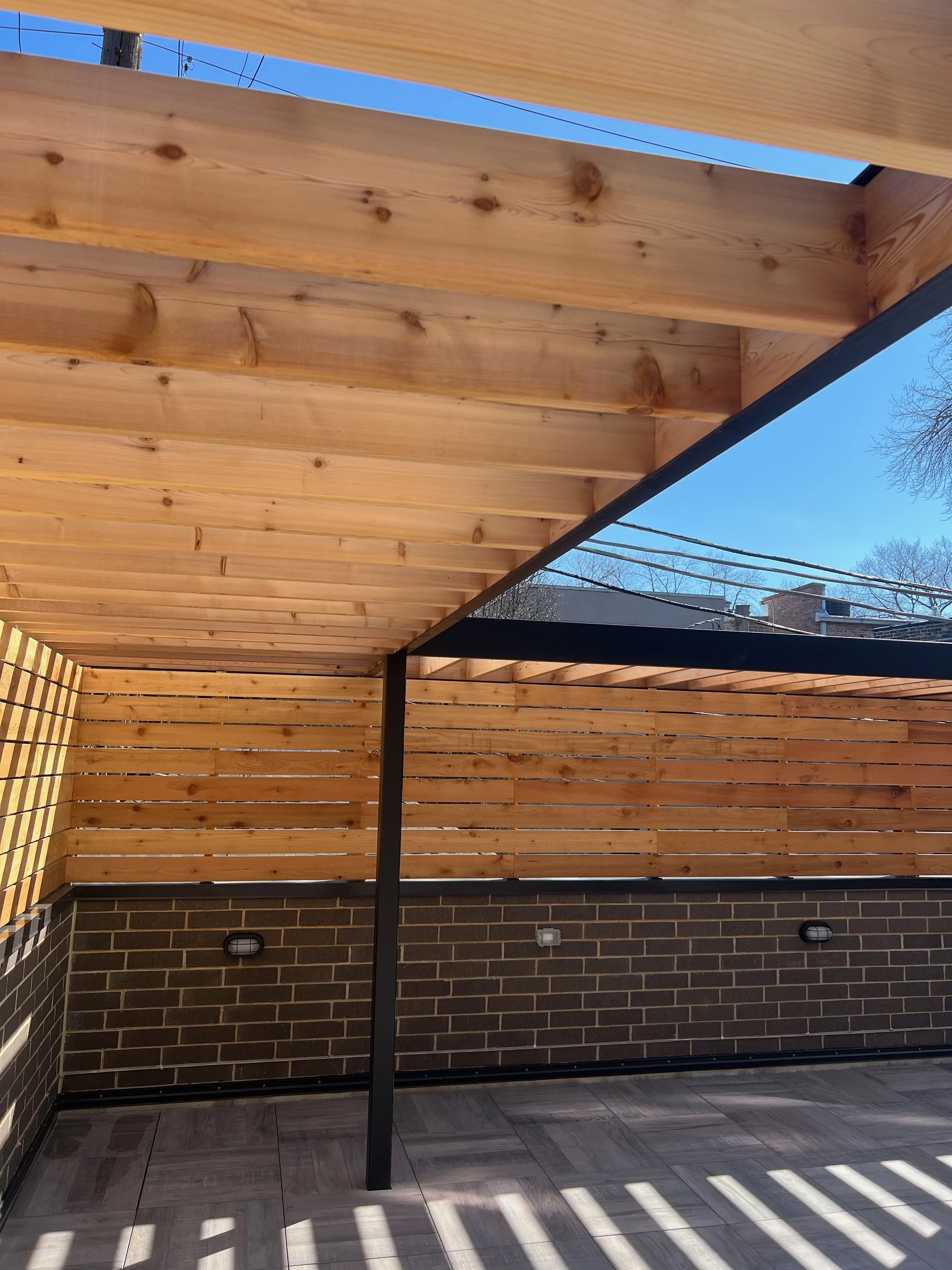 A newly constructed wooden patio or deck area with vertical slat fencing, brick wall, and tile flooring, under a roof with a black metal support beam, in a sunny outdoor setting.
