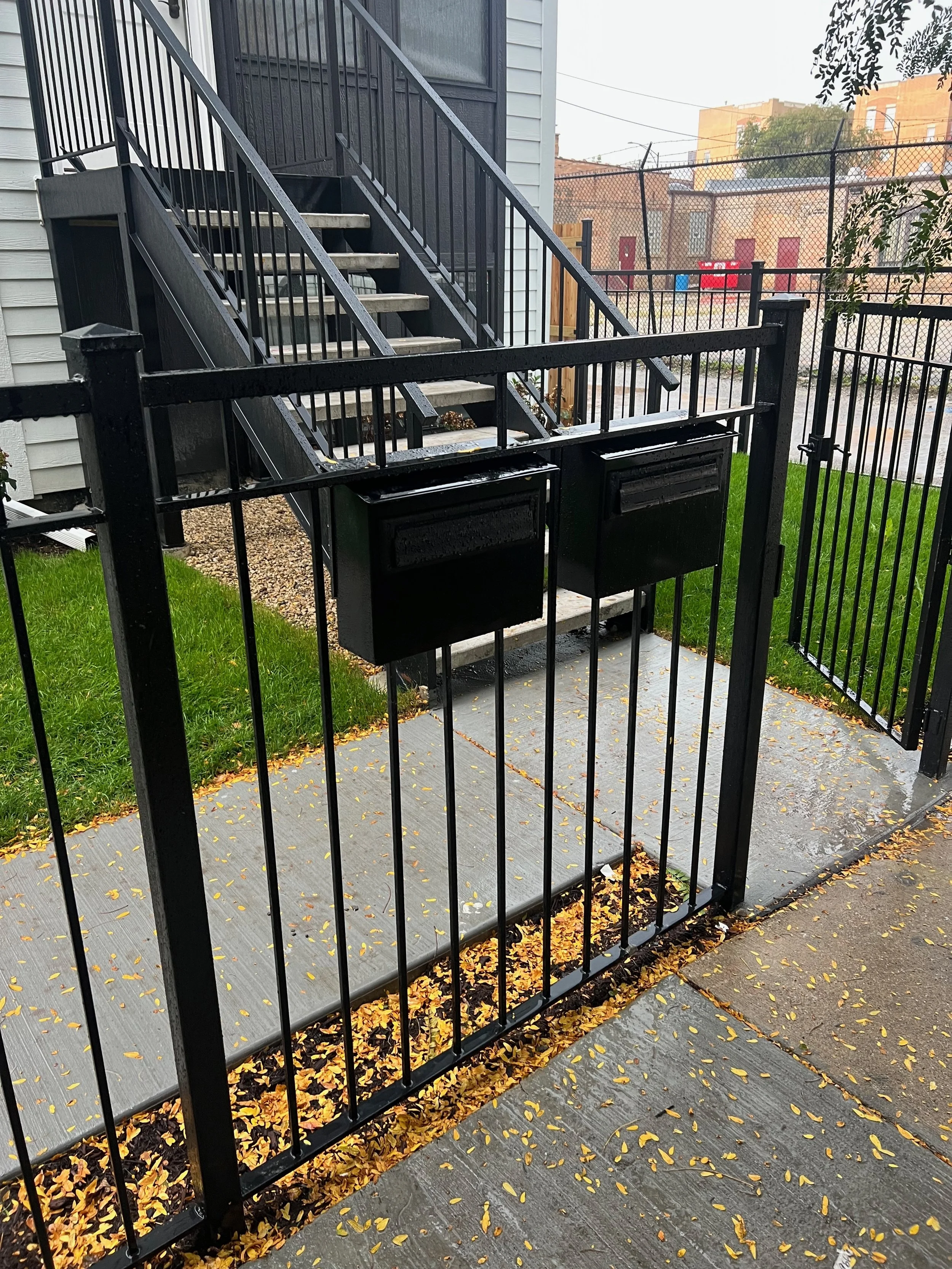 Rainy day view of black metal stairs leading to a closed door, with green grass, fallen yellow leaves, a black metal fence, and a cityscape with orange brick buildings in the background.