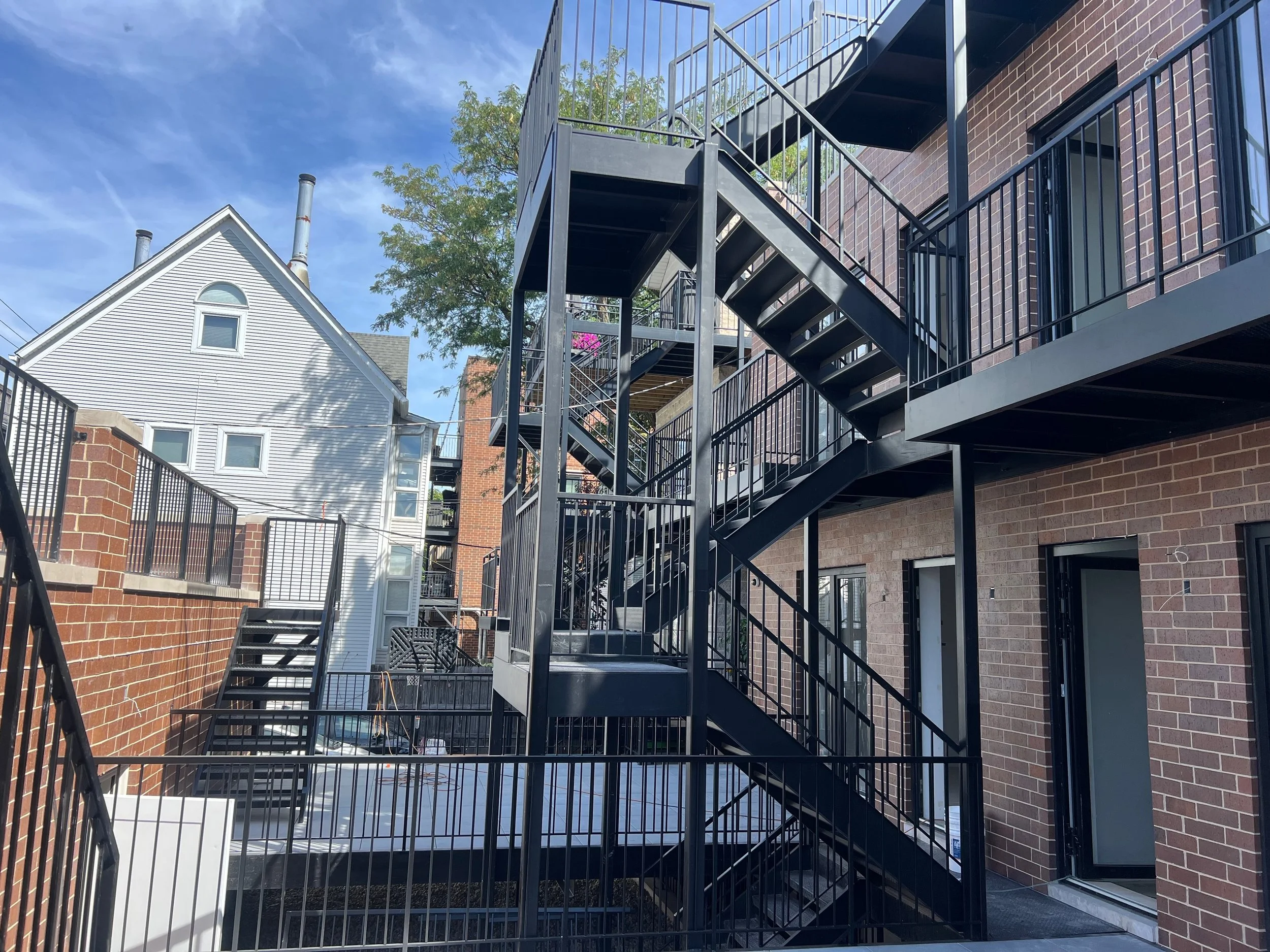 Black metal outdoor staircase with landings and railings attached to a brick building, with a neighboring white house with chimneys and a tree in the background.