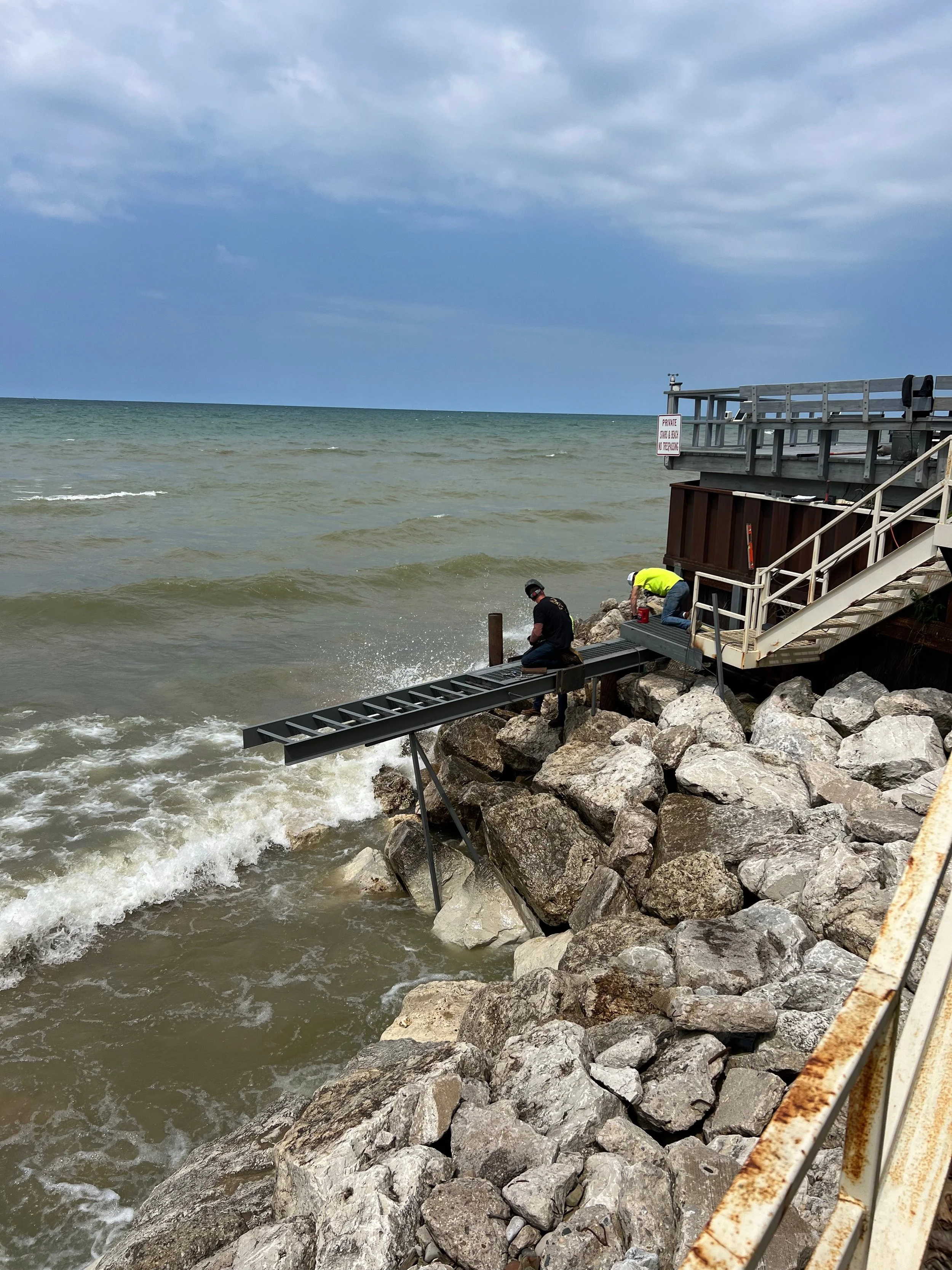 Two workers are installing rocks along the shoreline with construction equipment, including a metal ramp, under a cloudy sky, near a wooden pier with a sign that says "Private Shore & Beach Do Not Cross."