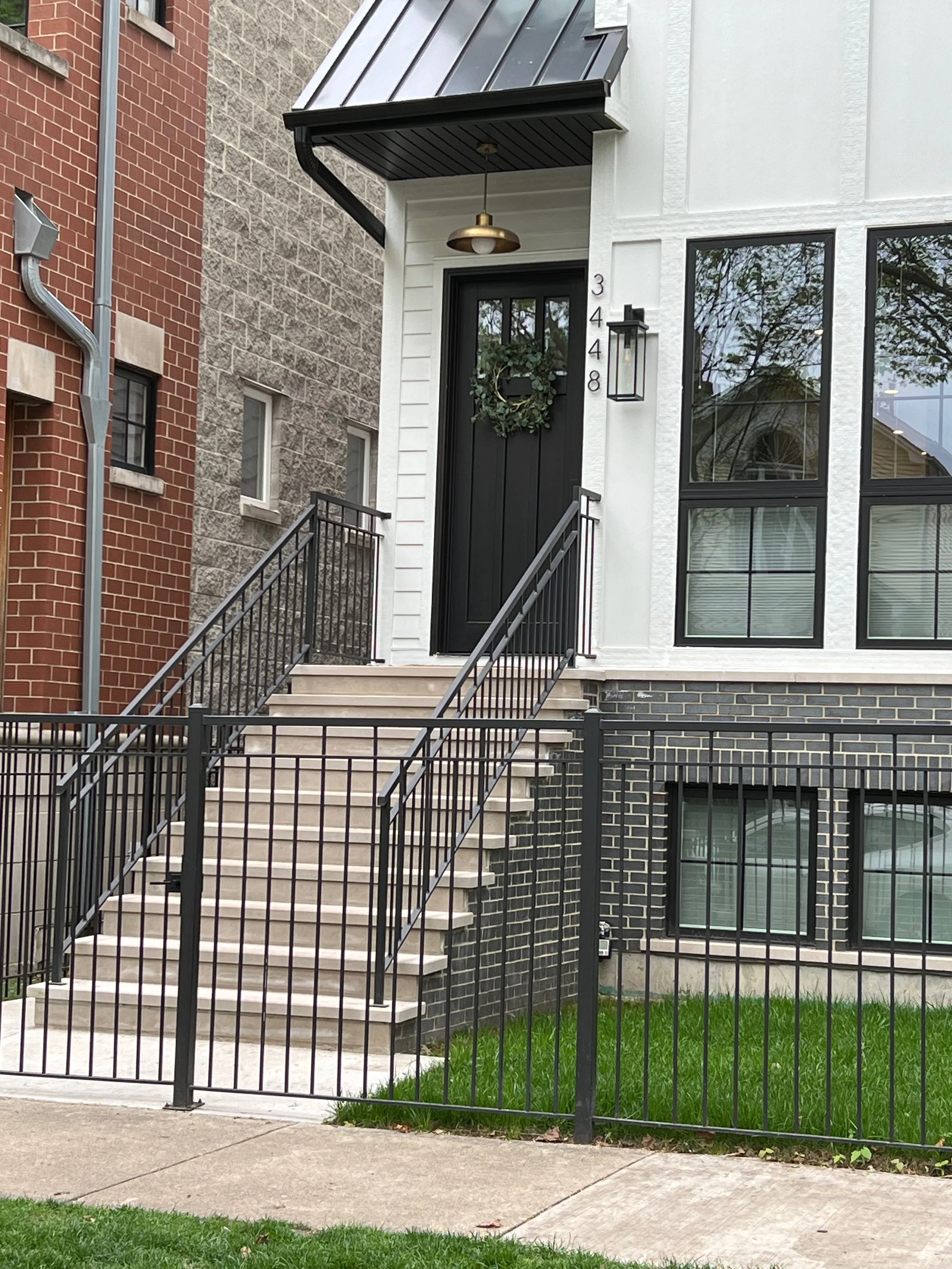 Front entrance of a modern home with black stairs, black door with a wreath, and house number 3448, surrounded by a black metal fence and a small grassy lawn. Adjacent buildings include a brick building on the left and a stone and white siding buildi
