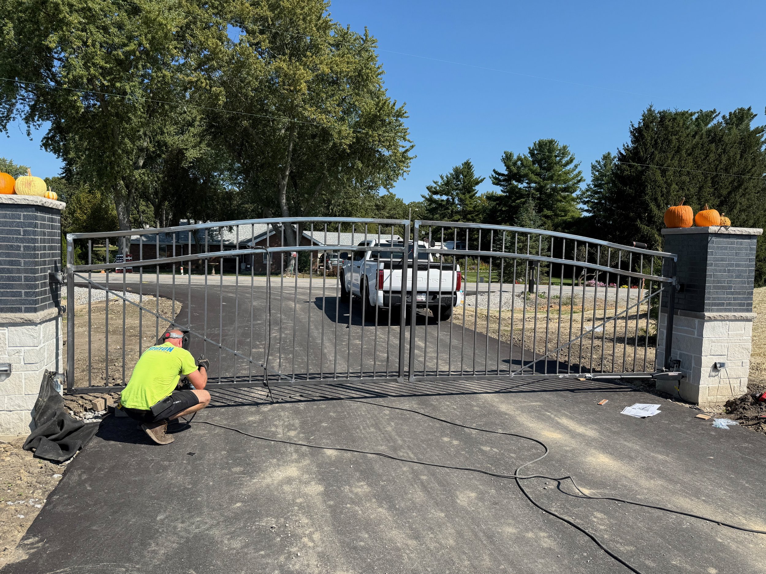Man installing or repairing a metal gate in a driveway, with pumpkins on brick pillars, large trees, and parked vehicles in the background on a clear day.