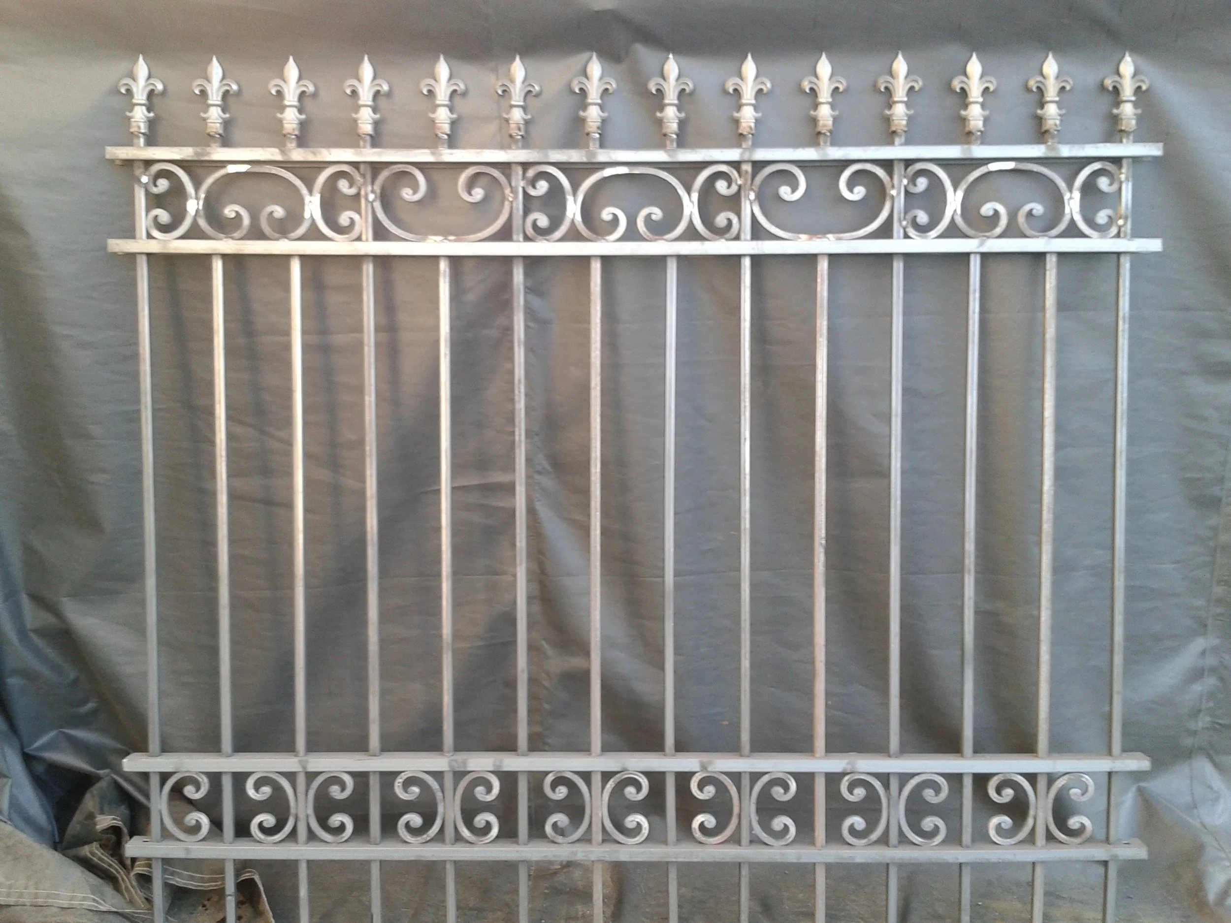 A decorative white iron fence with fleur-de-lis finials on top, featuring scrollwork details, against a plain background.