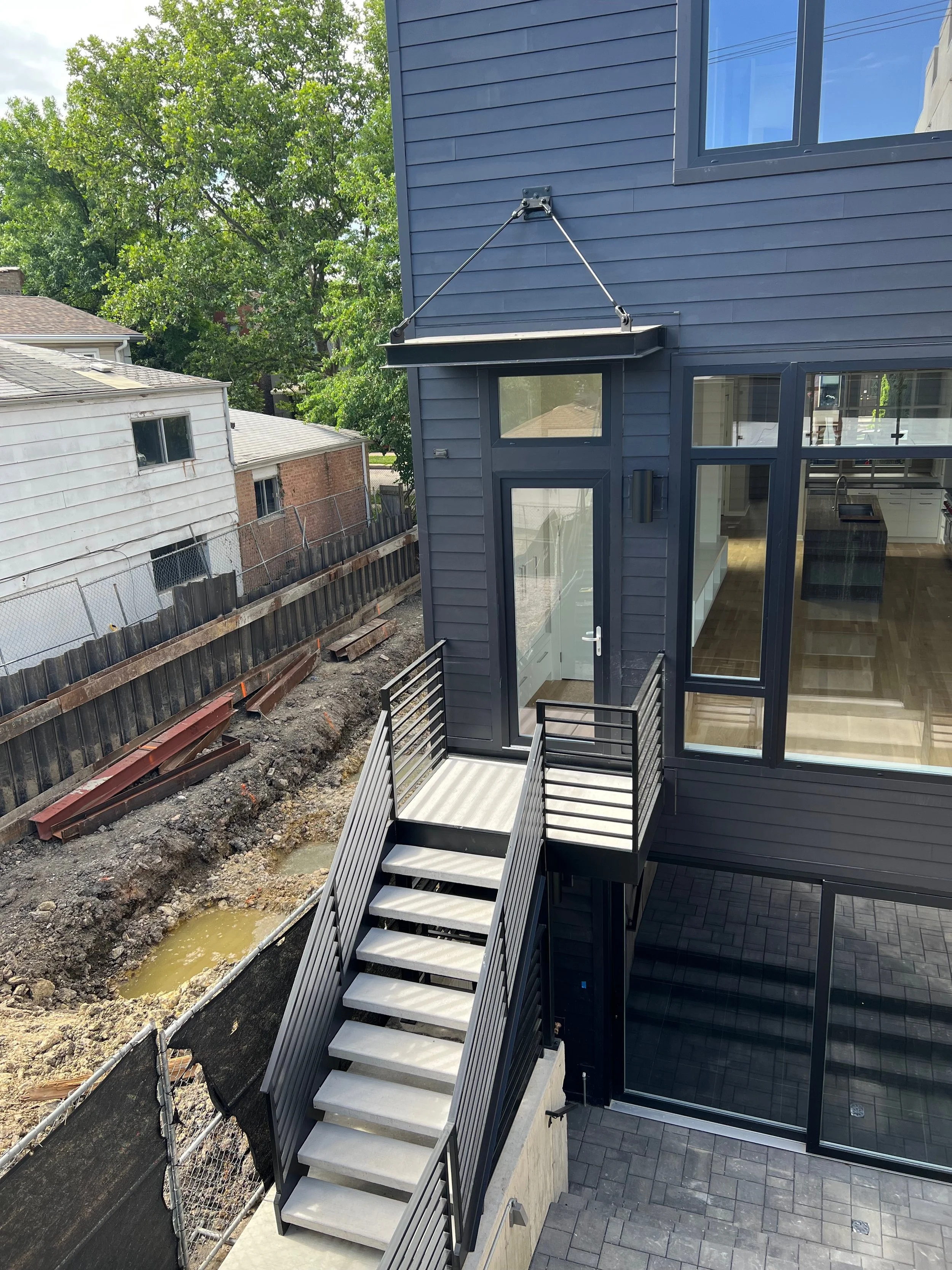 Exterior view of a modern house with dark blue siding and large windows. There is an outdoor staircase with metal railings leading to a door at an upper level, with a small deck at the top. The surrounding area has construction materials and an excav
