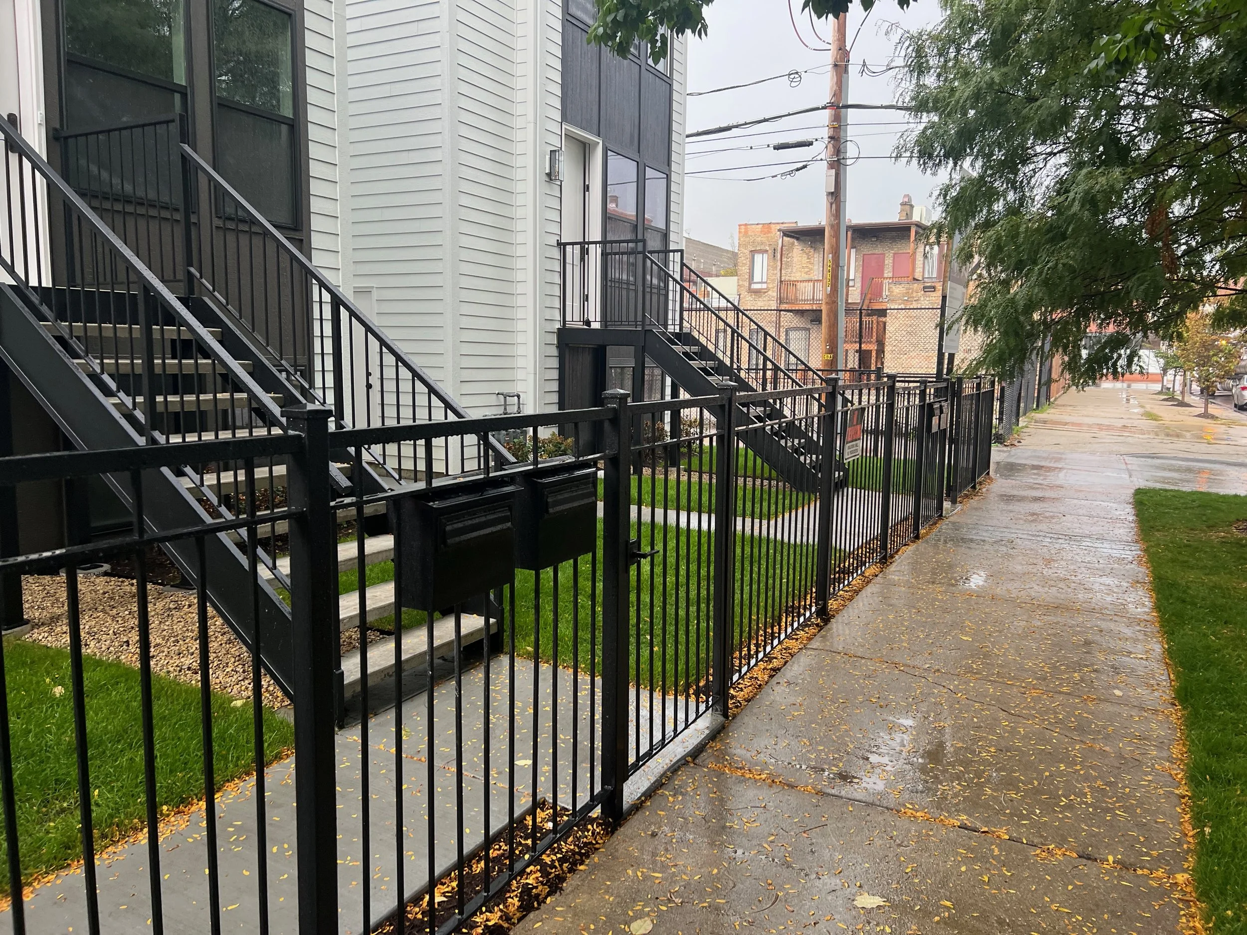 Sidewalk lined with trees, apartment building with external staircase and black metal fencing, rainy day with wet pavement and fallen leaves.