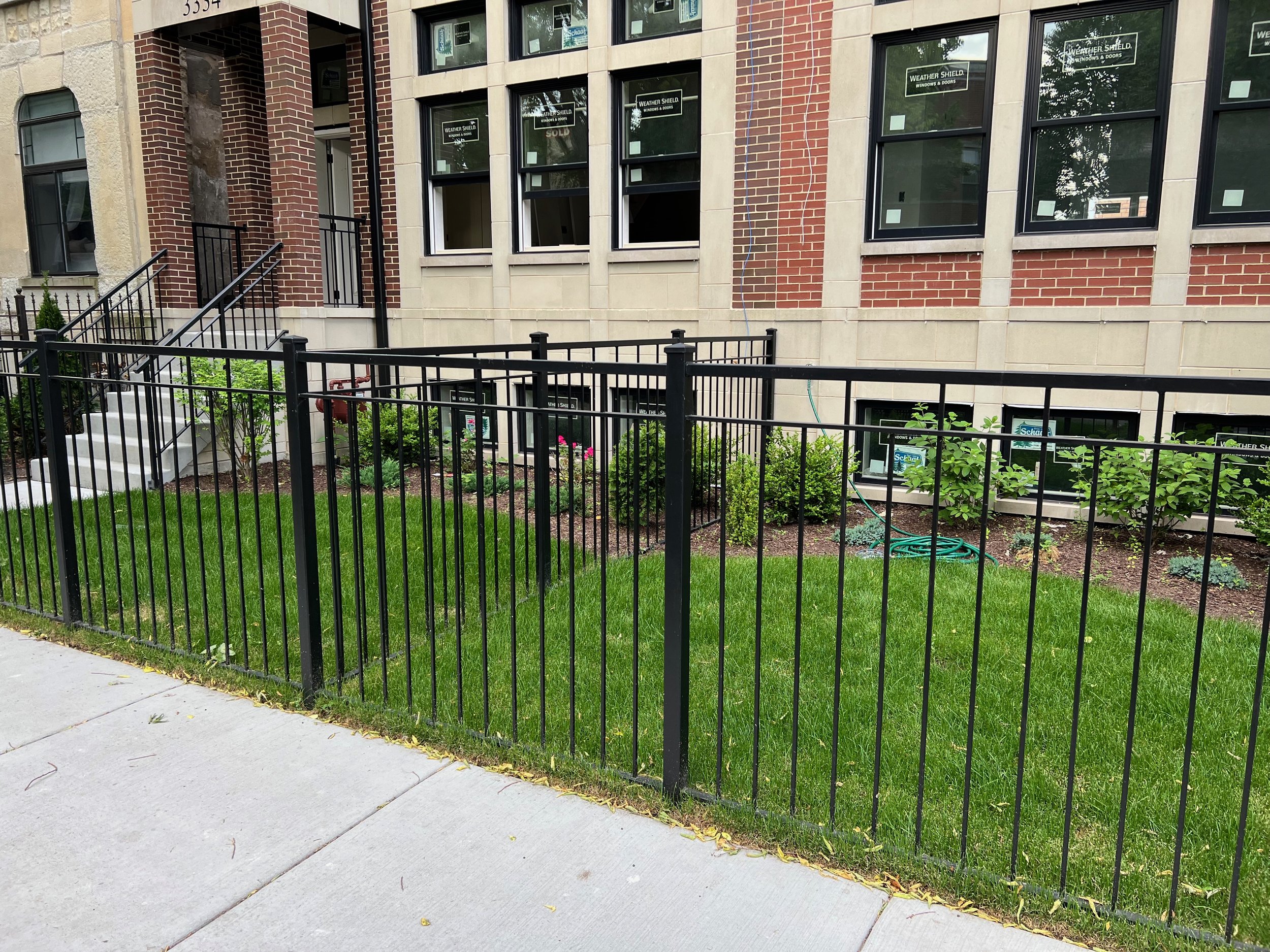 View of a residential building under construction with brick and stone exterior. There's a black metal fence in the foreground, a staircase with black railings leading to a door, and multiple windows, some of which are labeled as sold. There are some