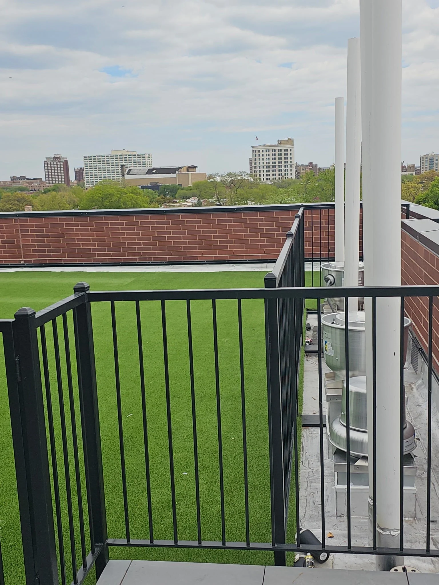 Balcony with green artificial turf, black metal railing, brick wall, and white pipes with equipment for utilities. City skyline with trees and tall buildings in the background under cloudy sky.