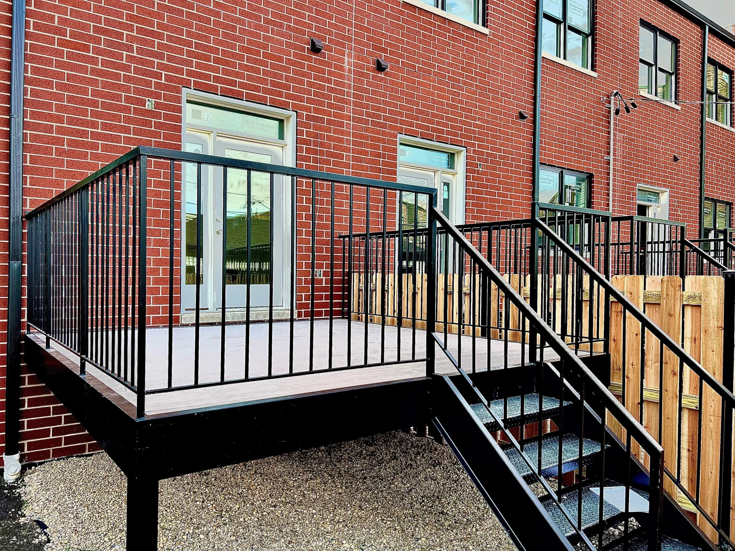 Apartment building with a red brick facade, black metal railing on a small porch, glass door with a molded frame, and multiple windows. Stairs with metal grating lead up to the porch, and wooden fencing surrounds the area.