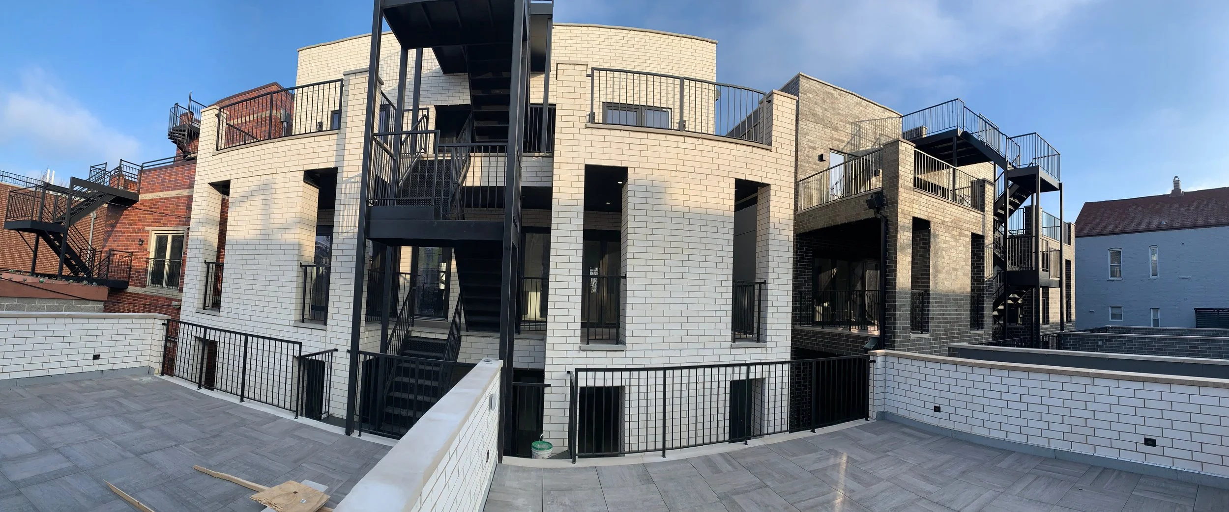 Modern multi-story building with white and gray brick facade, black fire escape staircases, and metal railings, with a rooftop patio and neighboring buildings visible under a partly cloudy sky.