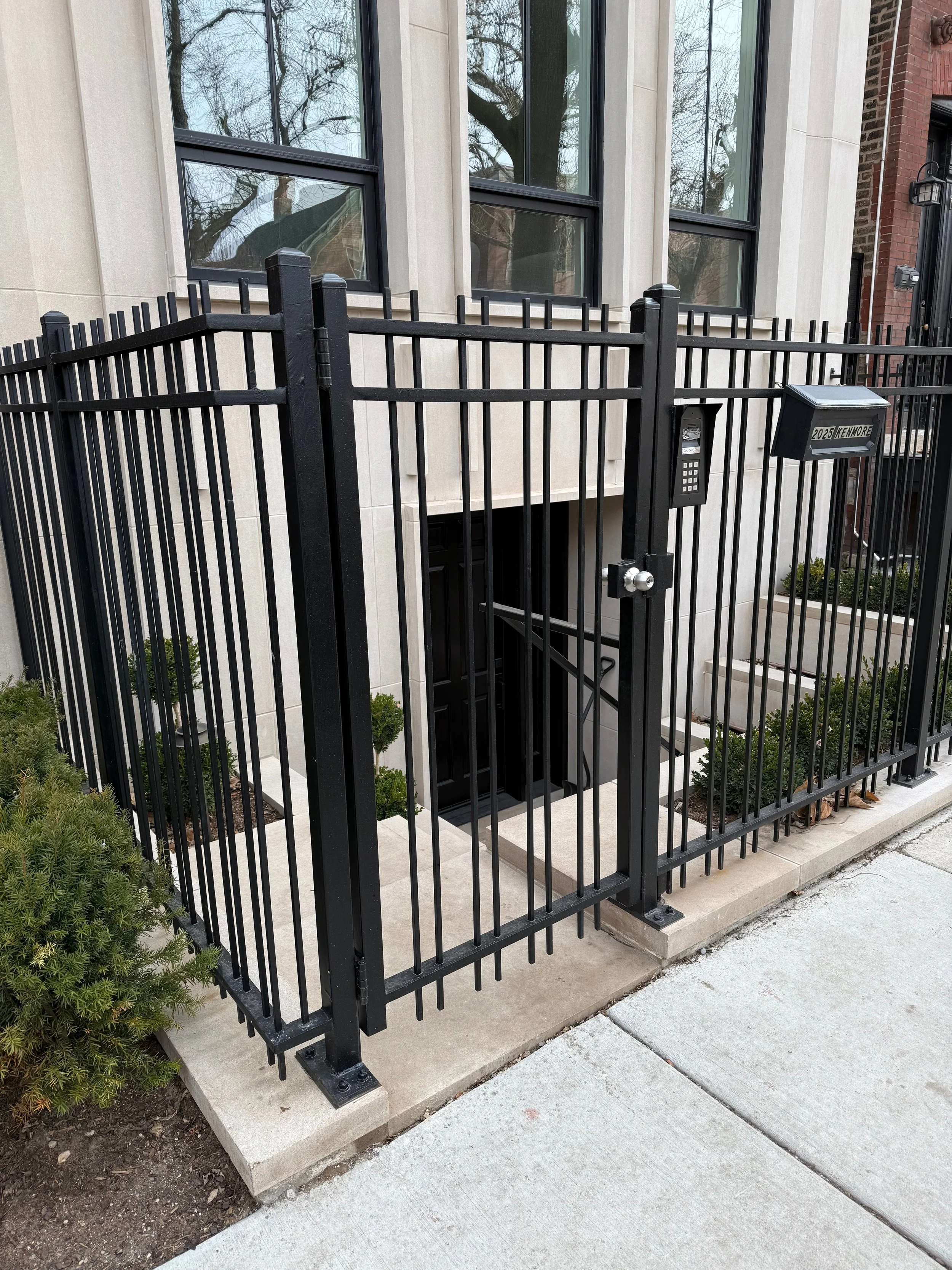Front entrance of a modern apartment building with glass windows, a black metal security gate, and a mail slot labeled '2025 Kenmore.' A keypad lock is attached to the gate, and there's a small garden with green bushes and concrete walkway.