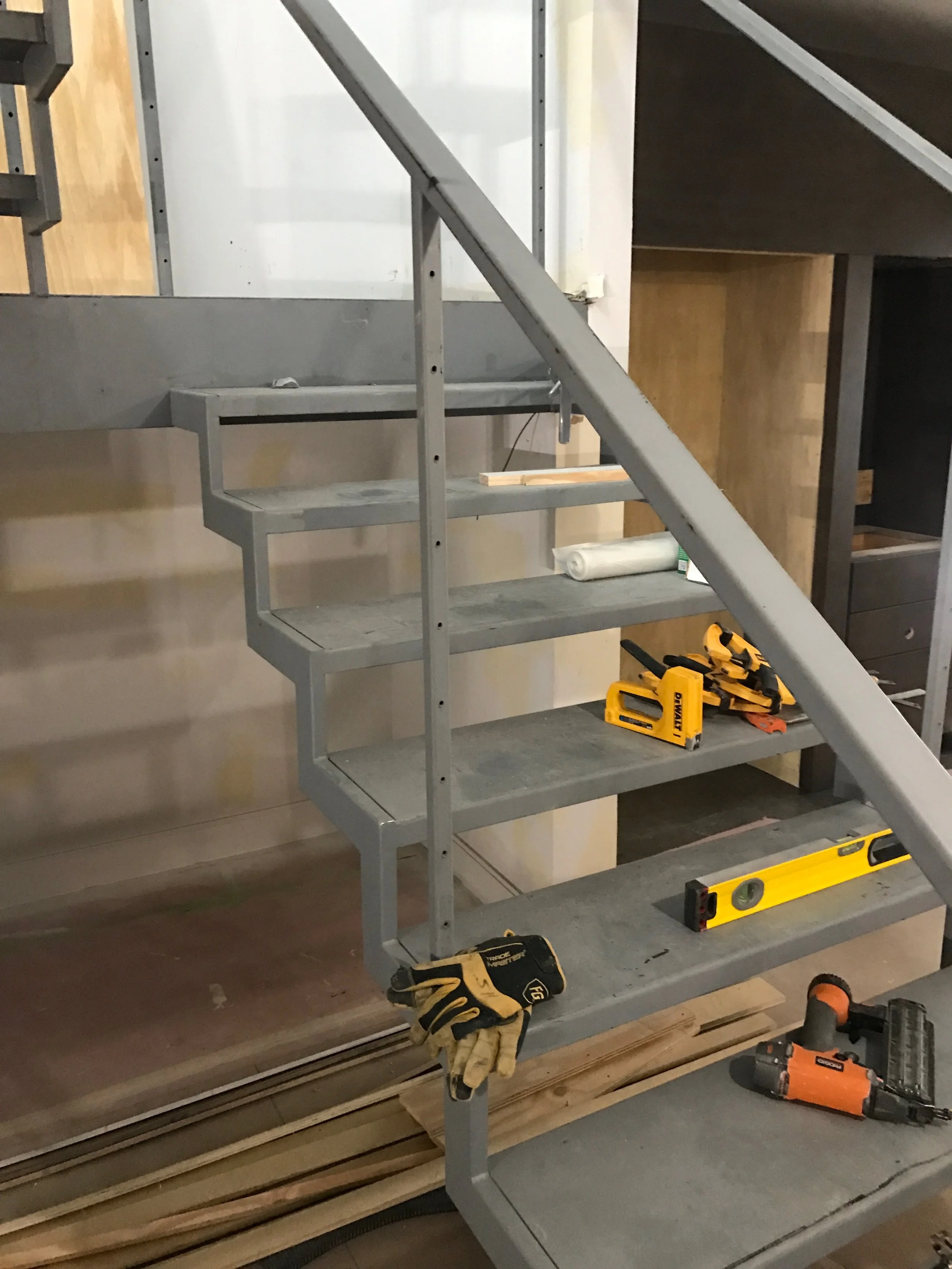 Construction site with metal shelving and tools. There are gloves, a level, a cordless drill, and a wood piece on the floor and shelves.