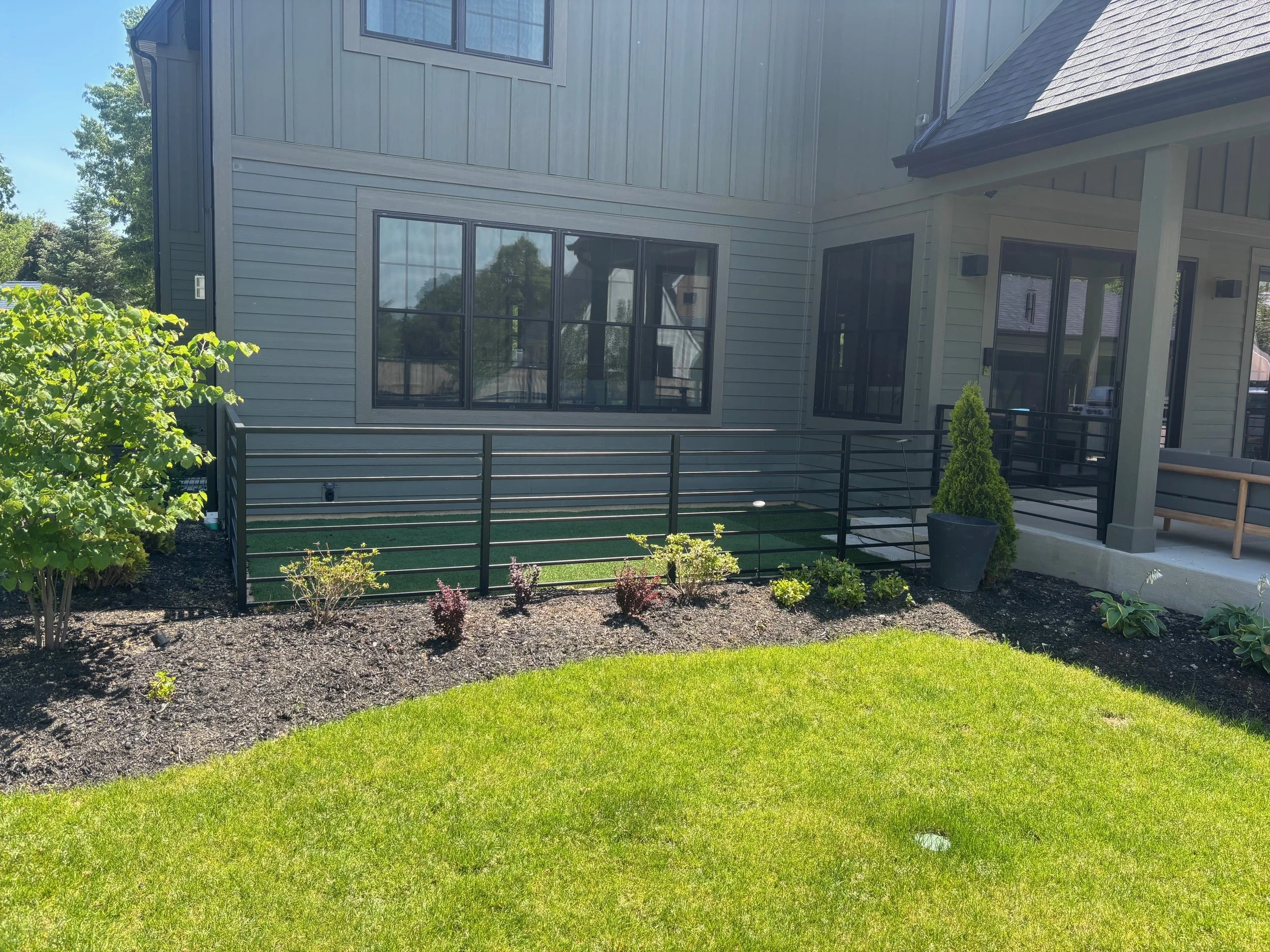 View of a backyard yard with a price of green grass, small shrubs, and a freshly mulched garden bed. The house has large black-framed windows and sliding glass doors, with a small porch area with a potted evergreen tree.