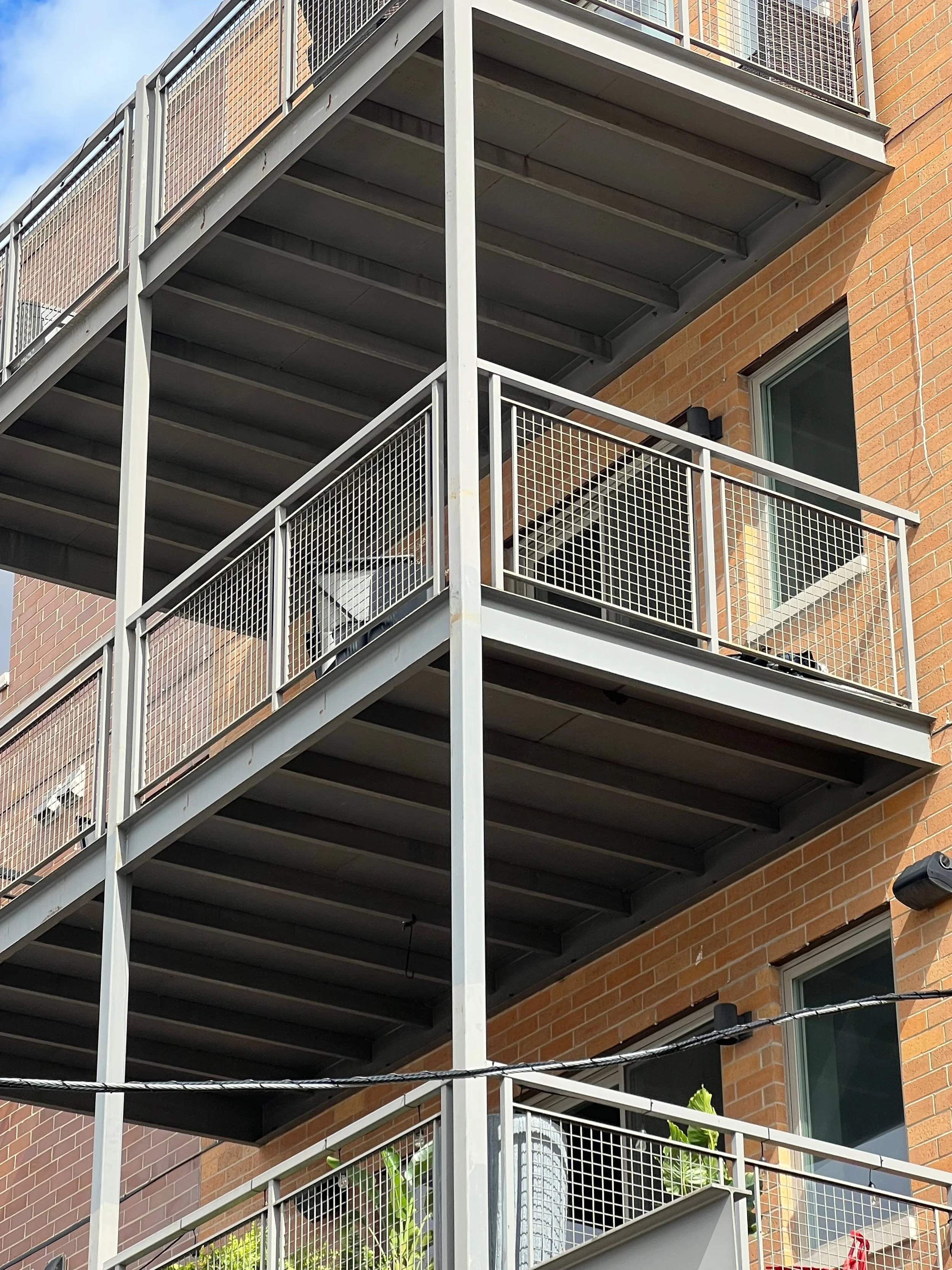 View of a modern apartment building with multiple floors and metal balconies featuring metal railings, brick walls, and windows with greenery below.