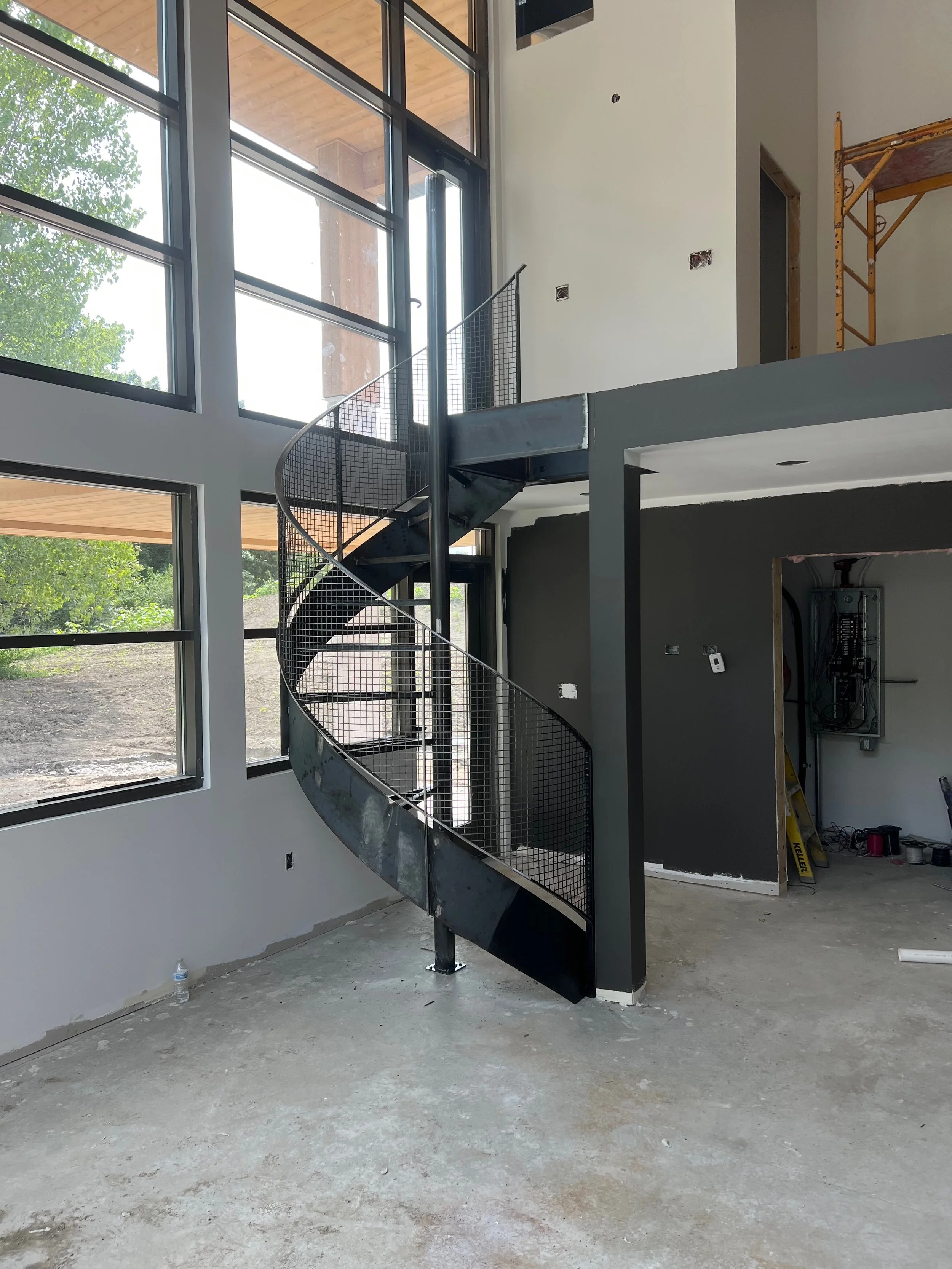 Interior under construction with large windows, a curved metal staircase with a black mesh railing, and unfinished walls and floor.