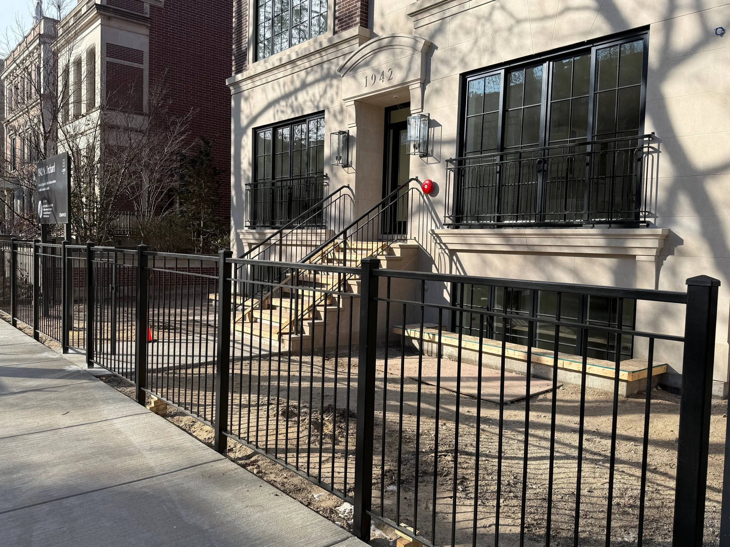 Construction area in front of a building with the year 1942 above the entrance, surrounded by a black metal fence, steps leading up to the entrance, and partially weathered wooden planks on the ground.