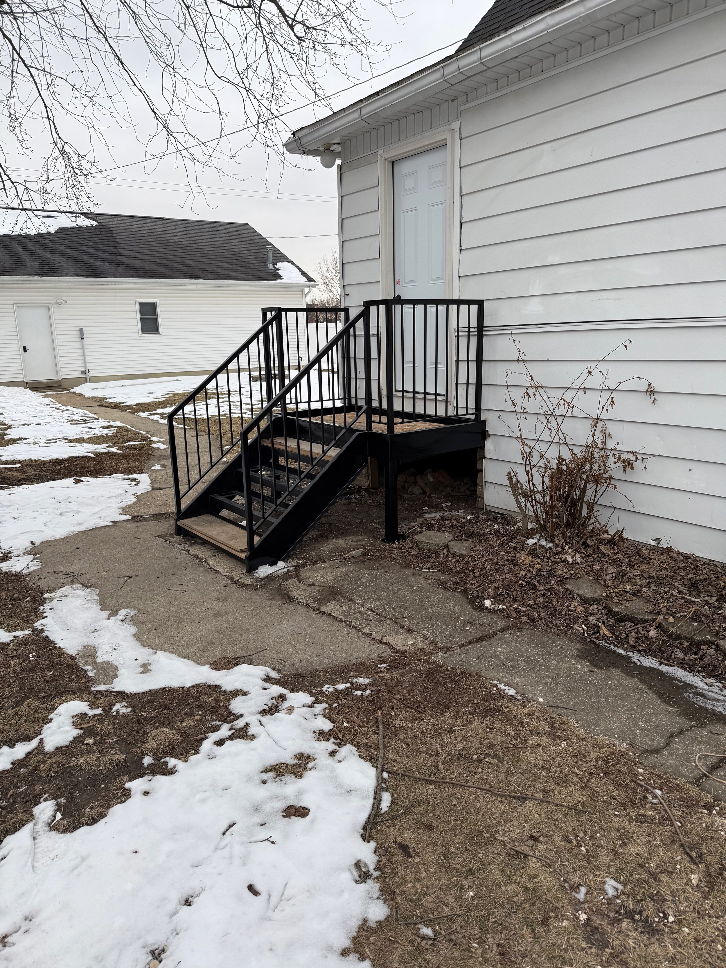 Exterior view of a house with a small staircase leading to a back door, with snow patches on the ground and leafless tree branches overhead.