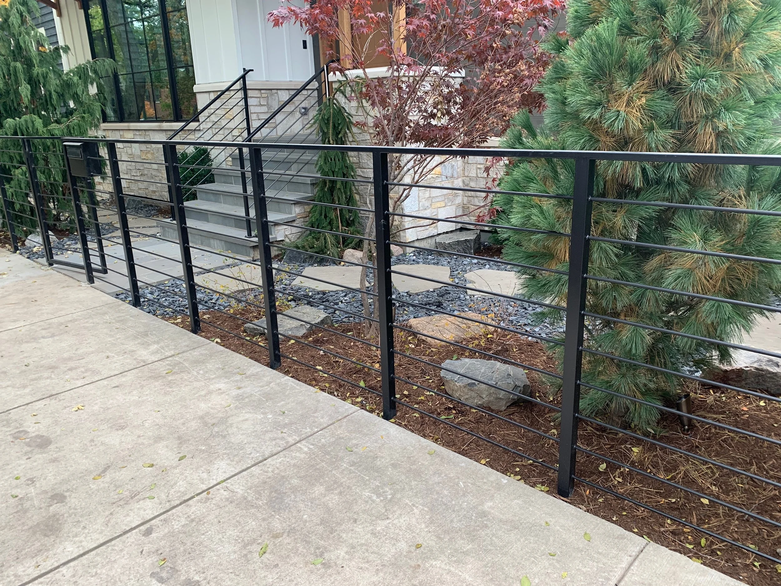 A front porch with a small staircase leading to a door, surrounded by a black metal fence, green bushes, and trees with red and green foliage.