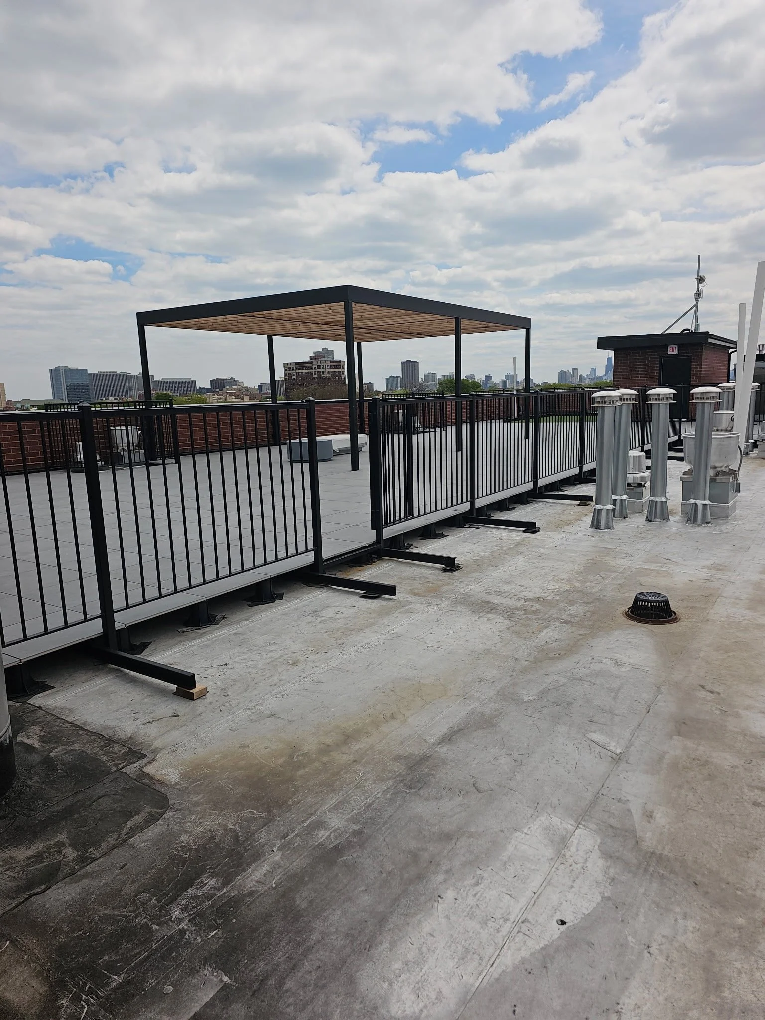 Rooftop with black metal railing, open structure with wooden roof, air vents, city skyline in the background, cloudy sky.