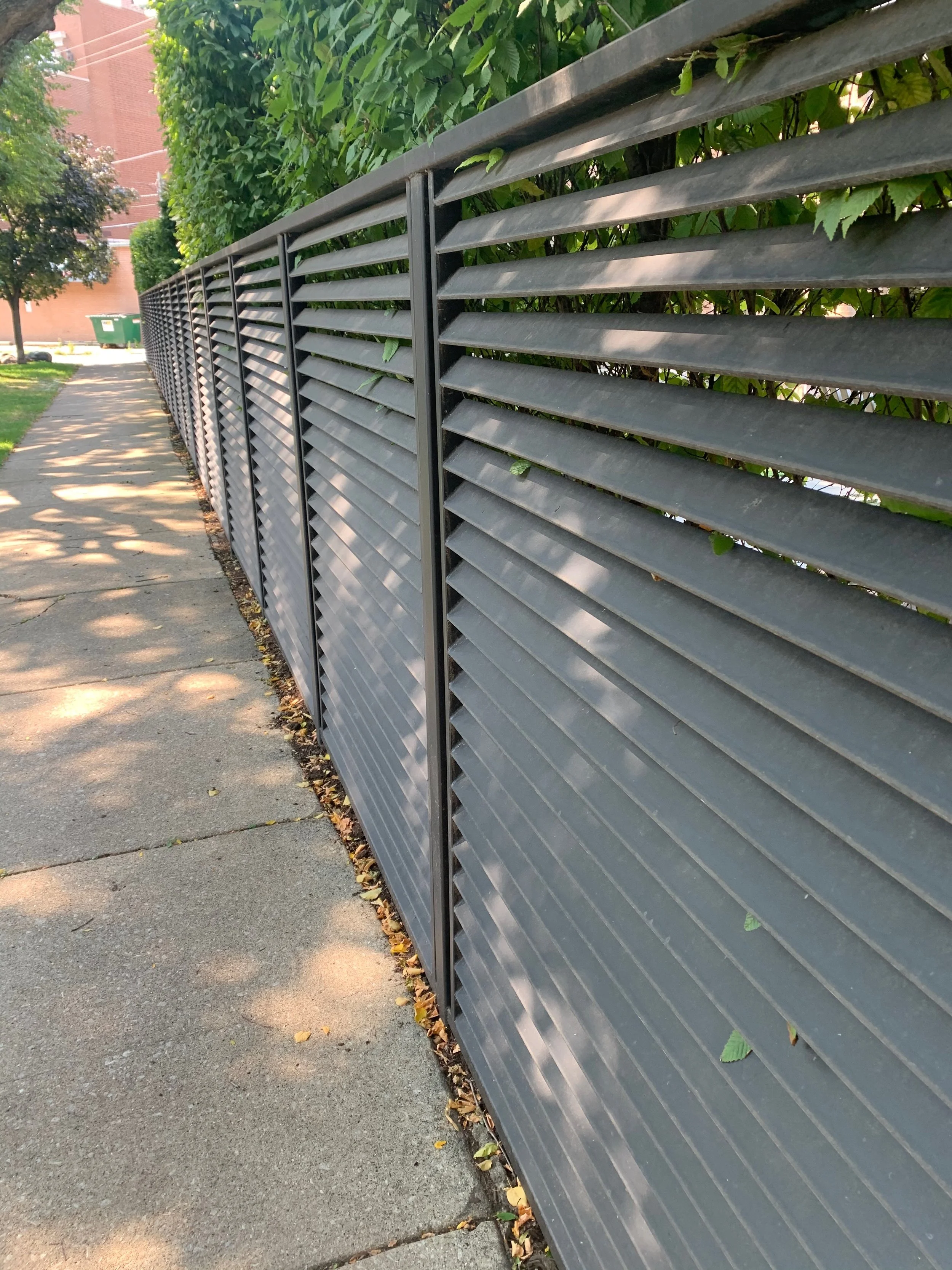 Side view of a metal fence with horizontal slats next to a sidewalk lined with fallen leaves, with a green hedge and trees in the background.