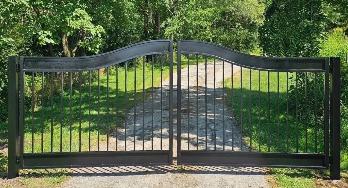 A black metal gate with a wavy top, open, leading to a dirt driveway surrounded by greenery and trees.