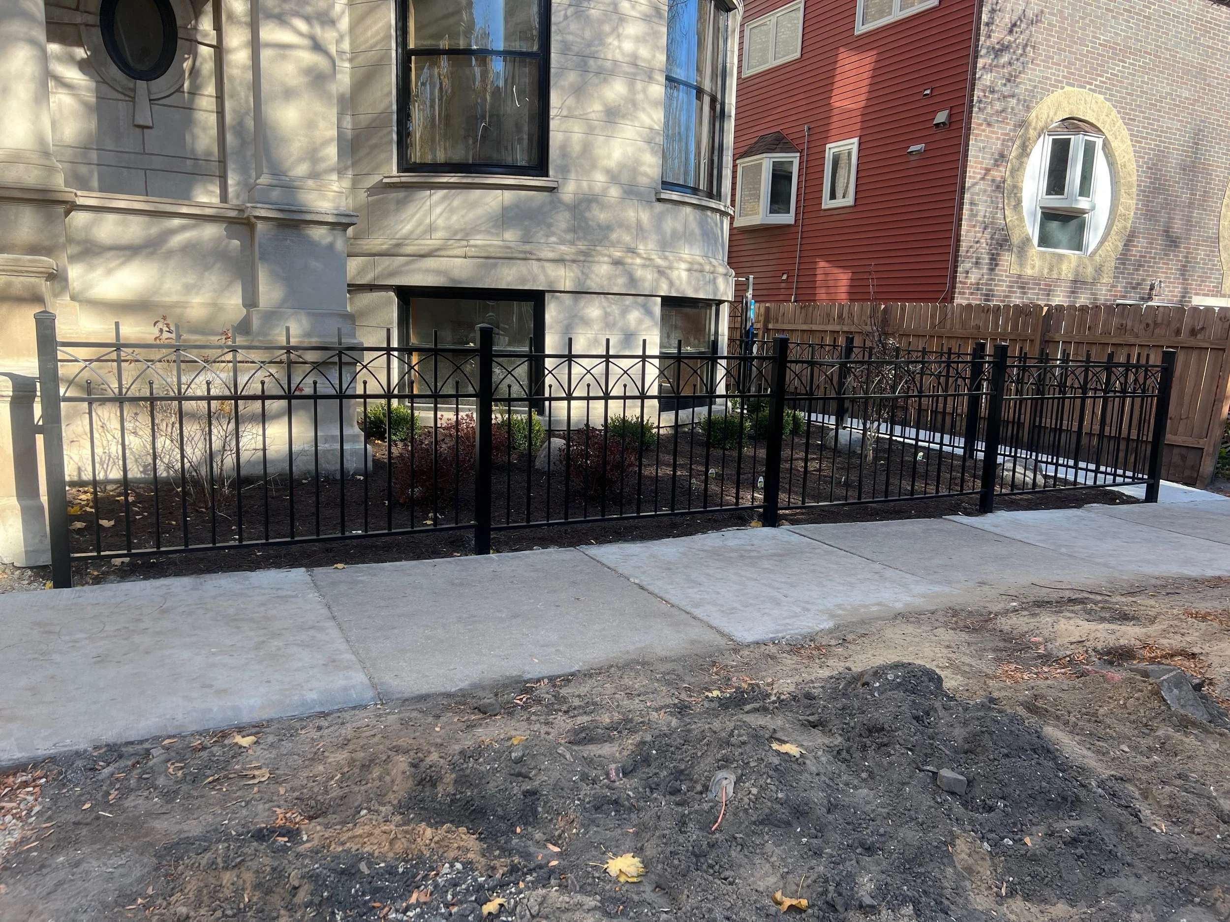 Side view of two residential buildings with small front yards separated by a black metal fence on a sidewalk, with underground landscaping under development.