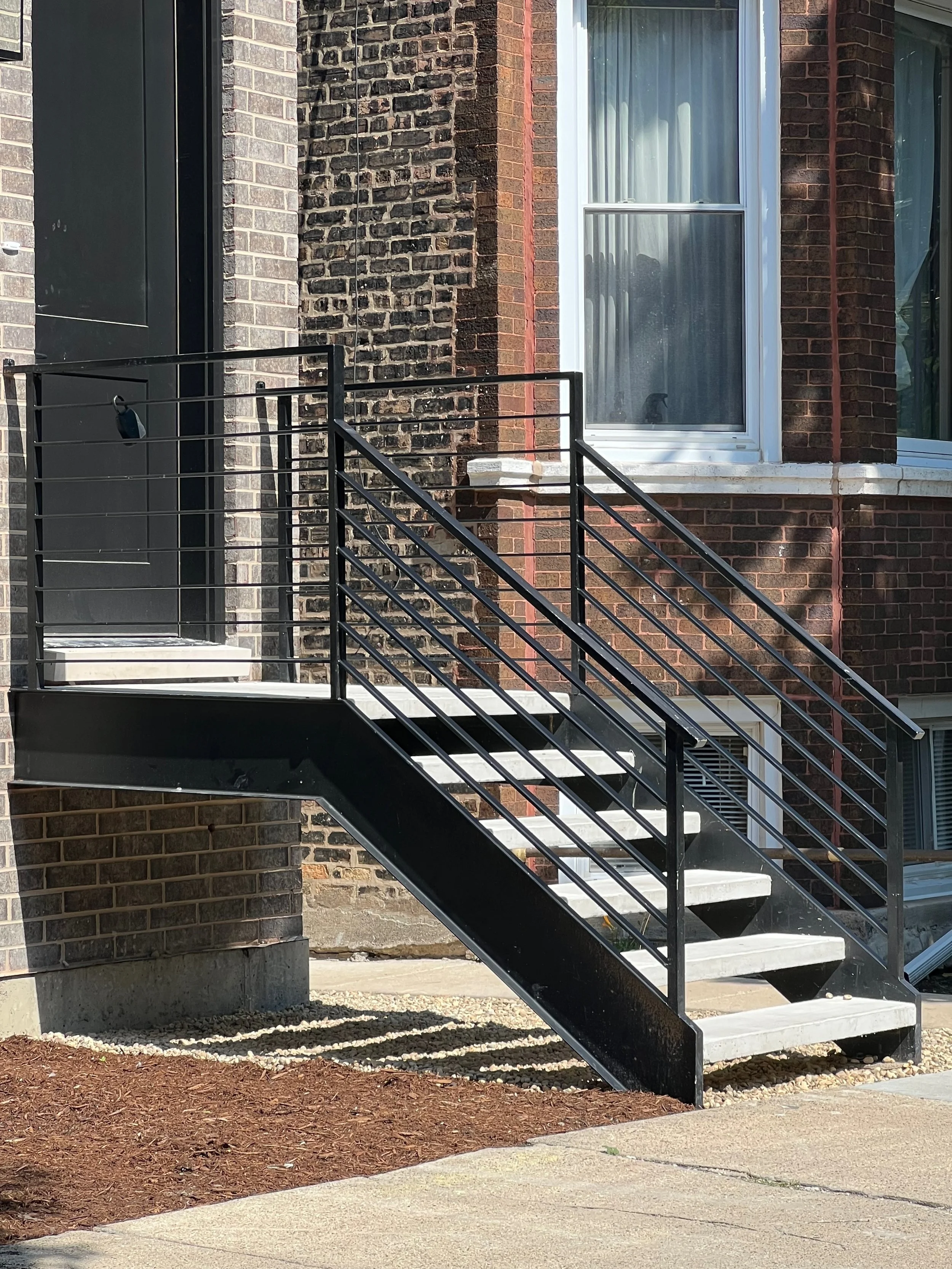 Exterior view of a brick building with a metal staircase leading up to a window and a door, with dirt and concrete ground in the foreground.