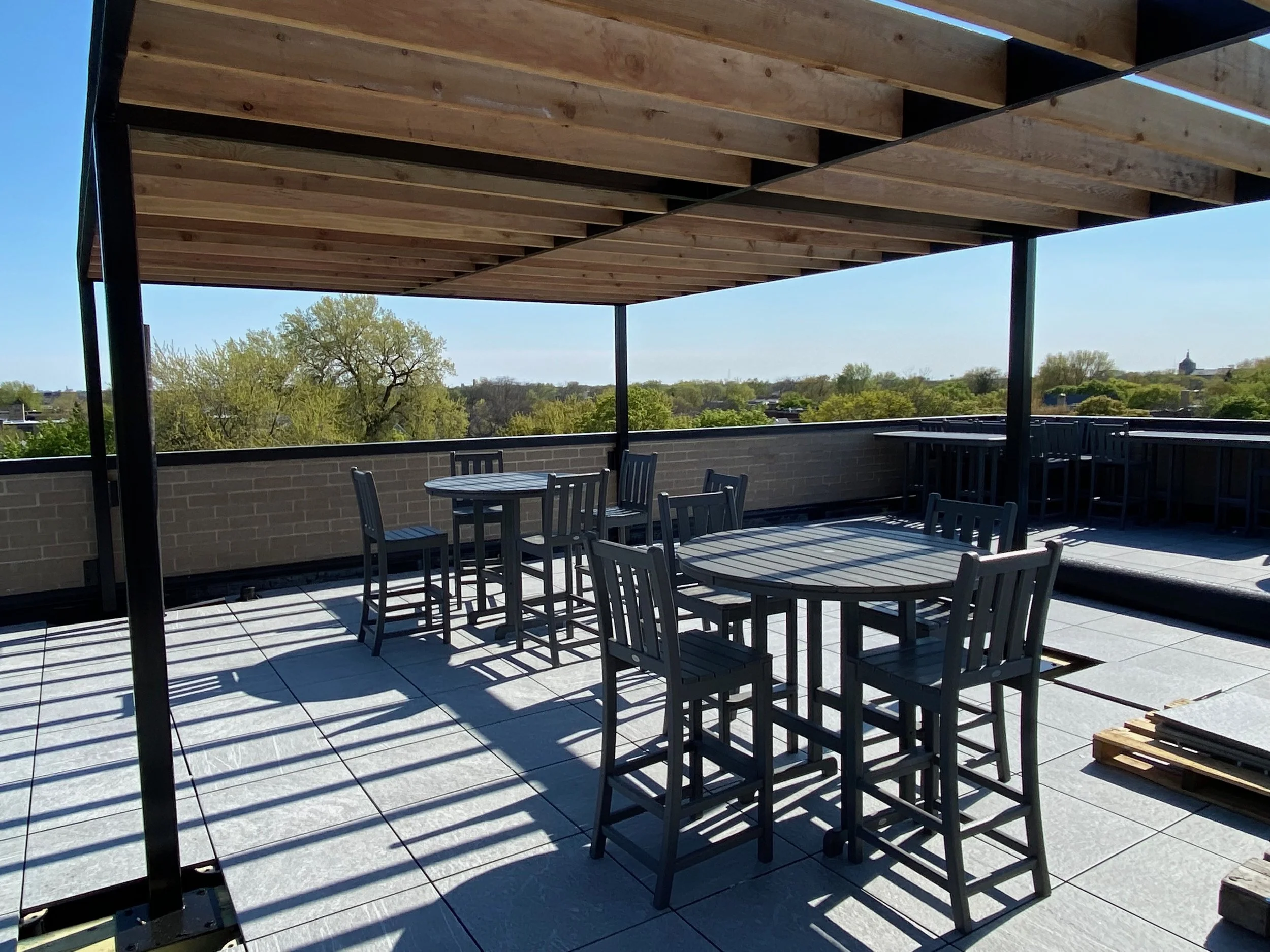 An outdoor rooftop patio with tiled flooring, a round table, several chairs, and a wooden pergola providing partial shade. In the background, there are trees and a cityscape with a church steeple visible.