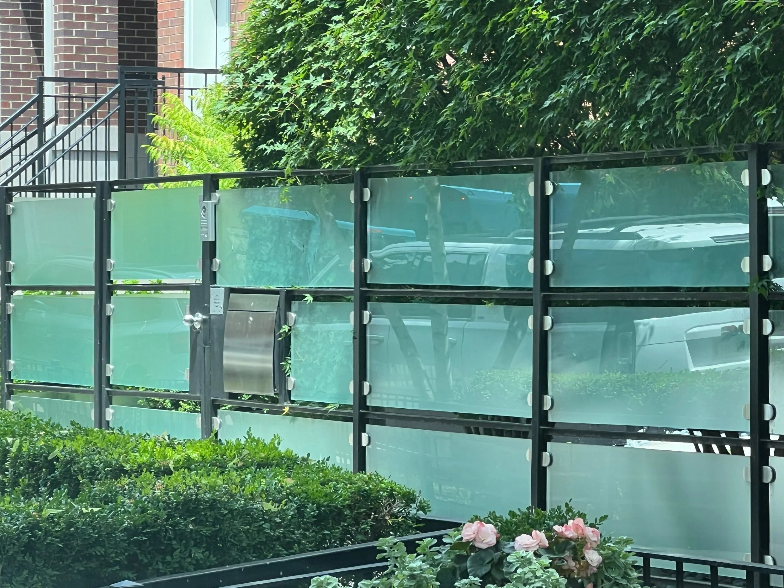 A black metal fence with glass panels surrounds a small garden with bushes and pink flowers. In the background, there is a red brick building with stairs and a balcony covered with green foliage.