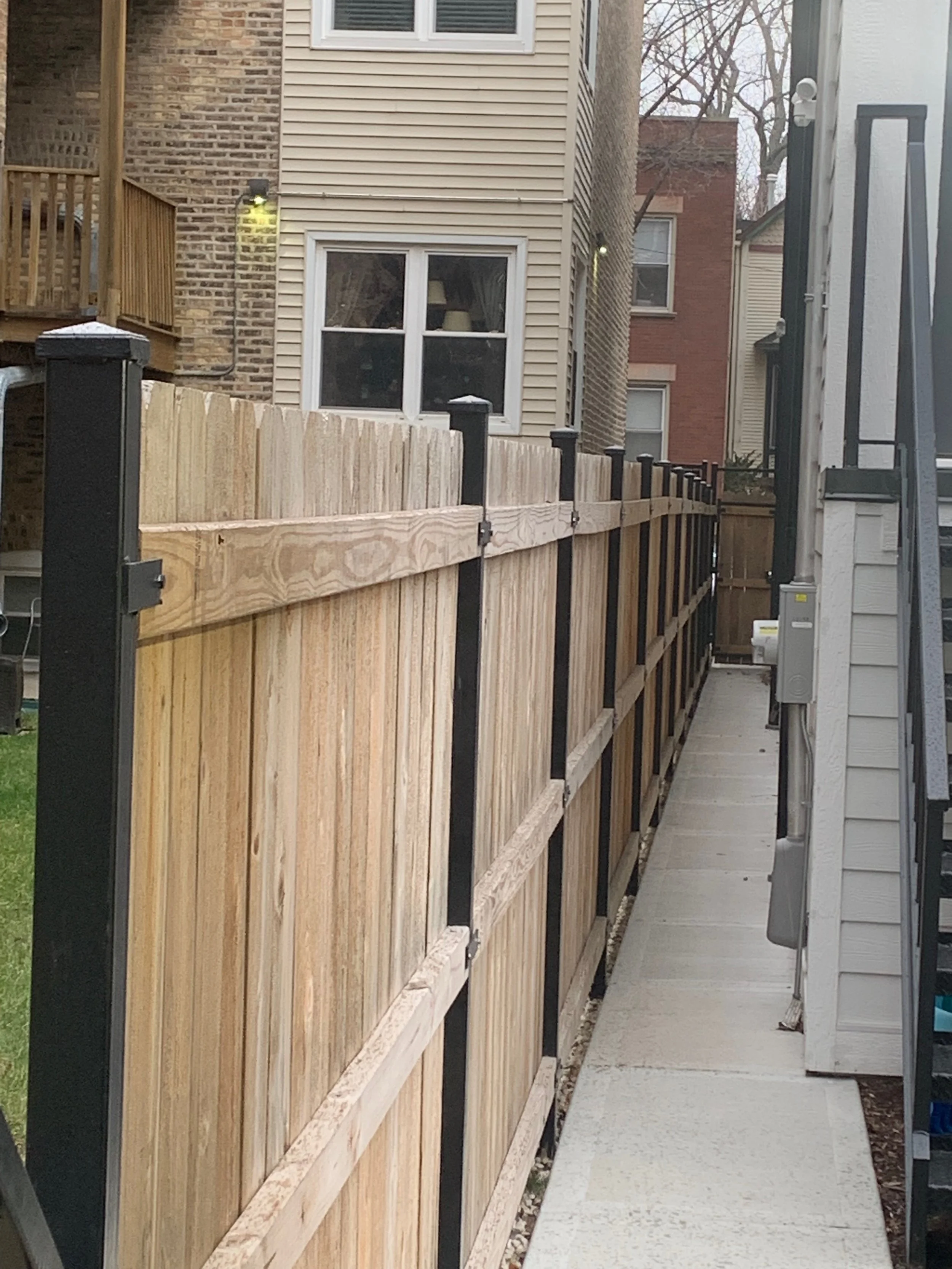 A side yard between residential houses, showing a wooden fence and concrete pathway, with brick and siding house walls, two windows, and some trees in the background.