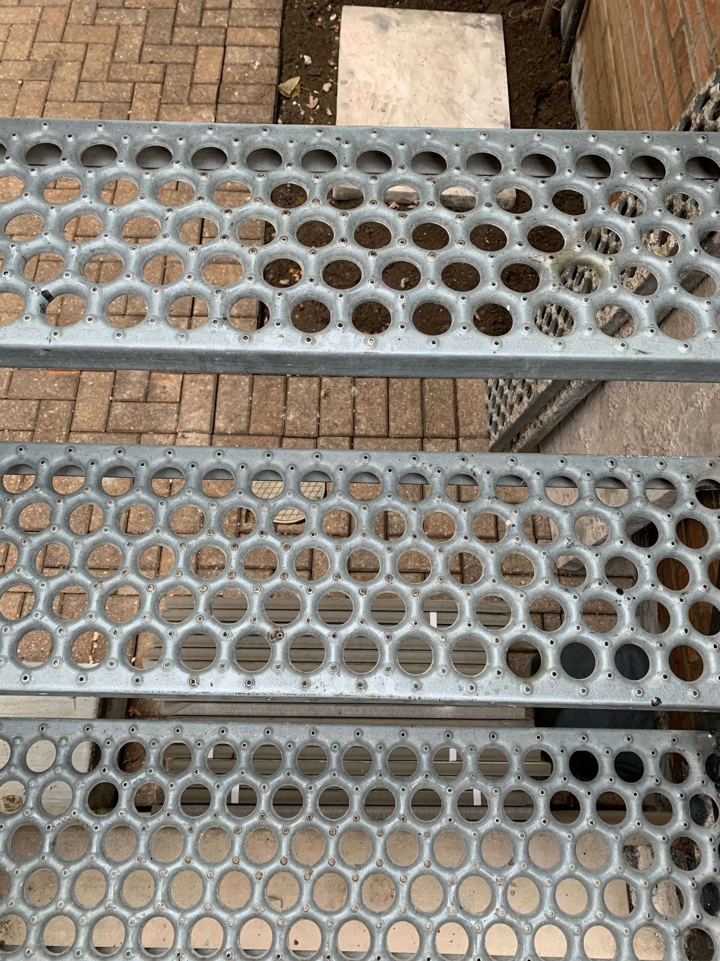 Close-up of a metal bench with circular hole patterns, situated outdoors on a brick and concrete paved area.