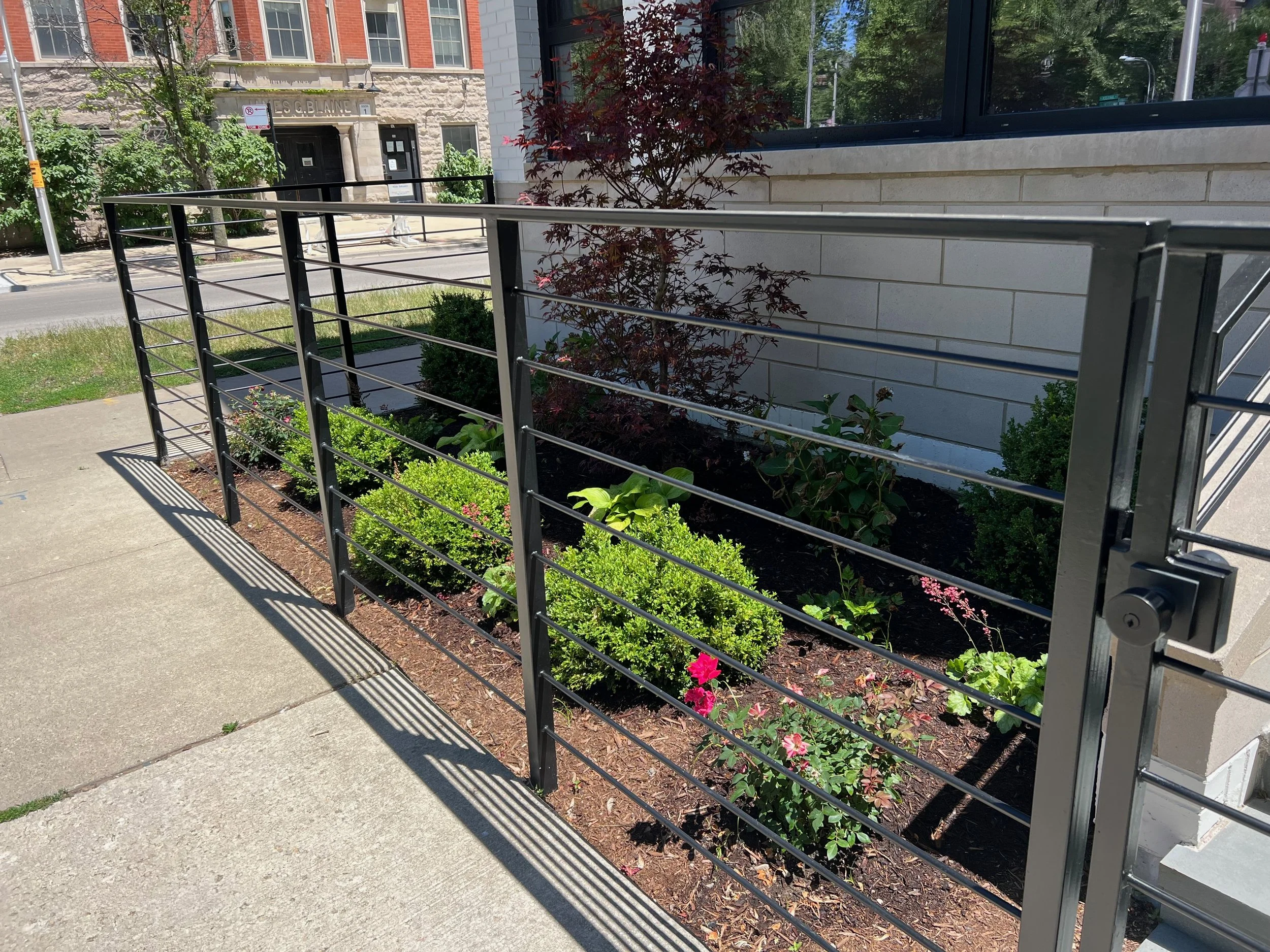 A small garden bed with bushes, pink flowers, and a red-leafed tree, situated next to a sidewalk and a building with black and white brick exterior.