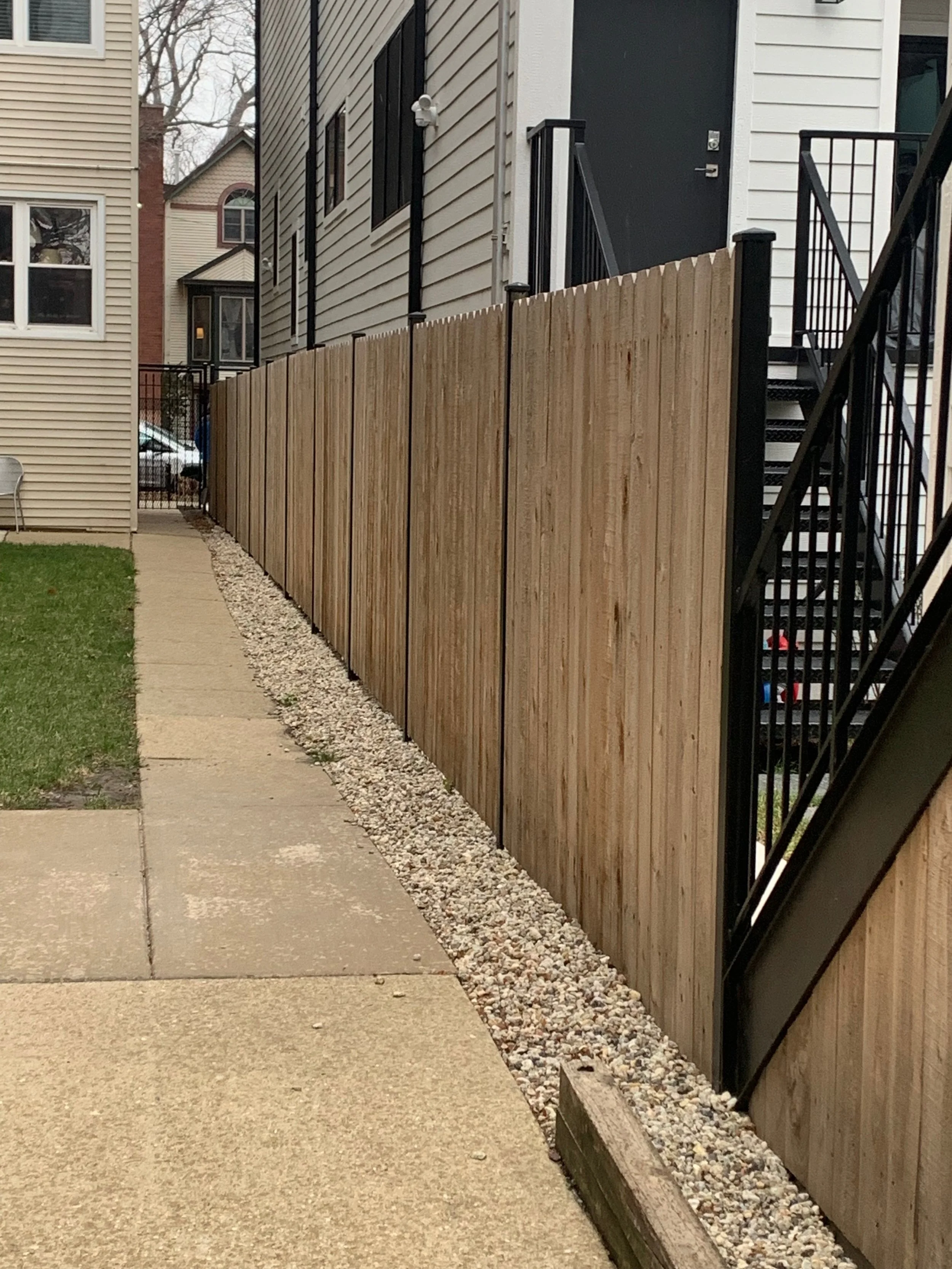 Side view of a residential outdoor area with a wooden fence along a walkway, neighboring houses, and stairs leading to a door.