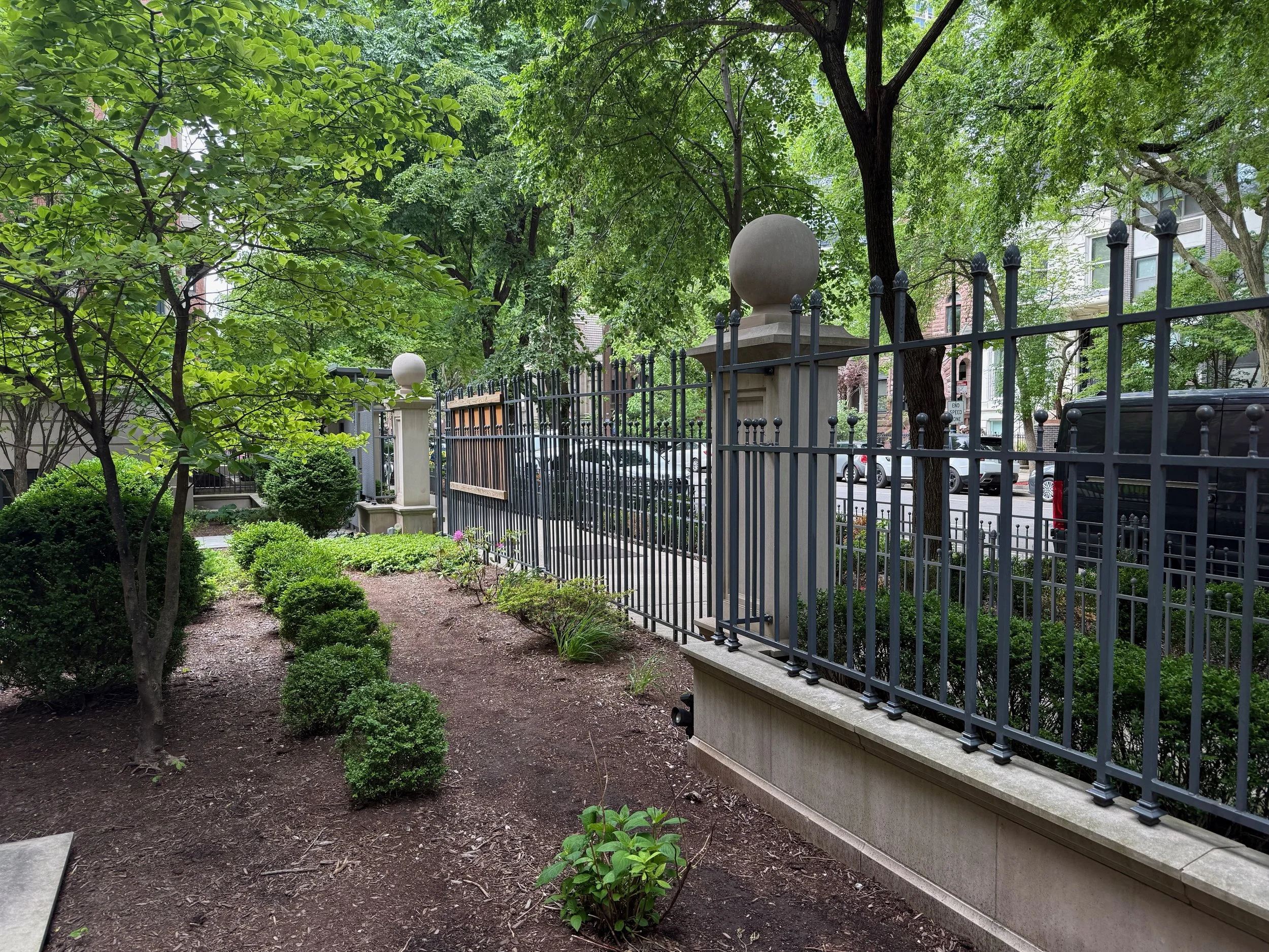 A fenced garden area with green trees, bushes, and a pathway. Large decorative stone spheres are mounted on the fence posts. Buildings are visible in the background.