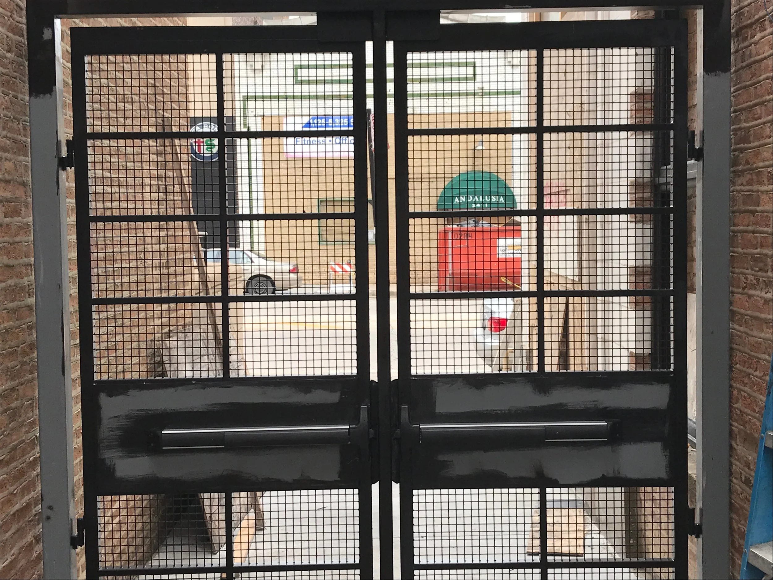 A gated stairway entrance with brick walls on either side, leading to a parking lot with cars and a building in the background. The gate has a mesh design with black framing.