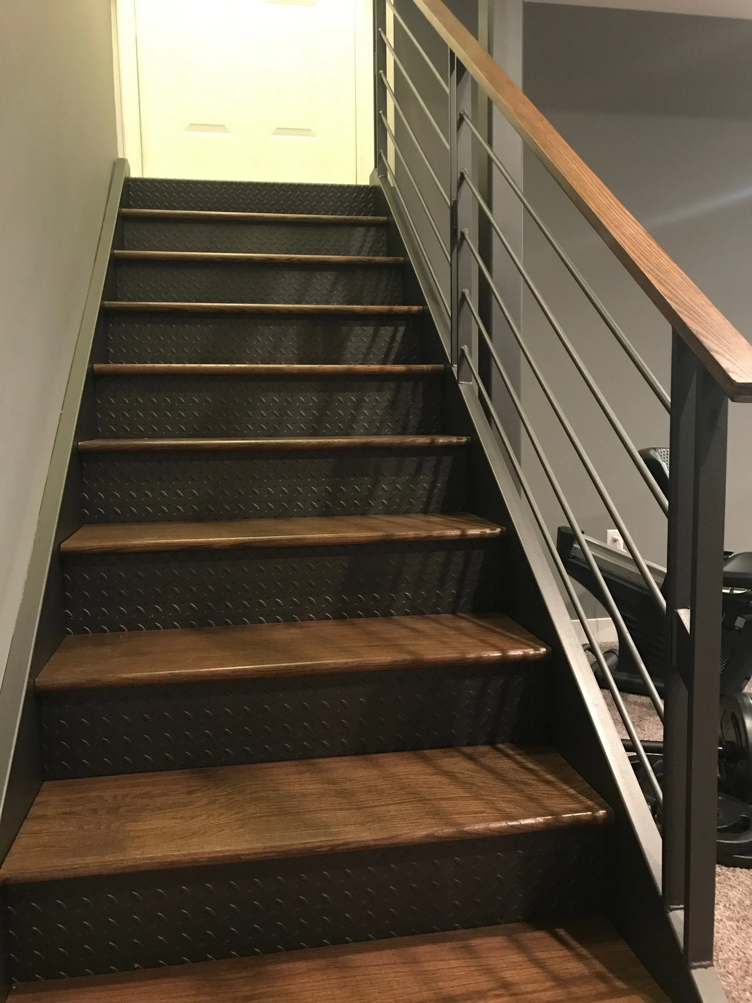 A staircase with metal framework, wooden steps, and a black textured riser. A metal handrail runs along the right side, and a white wall and door are visible in the background.