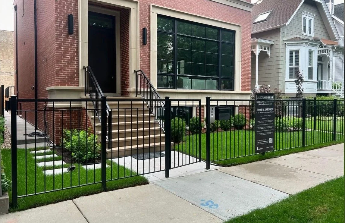 Newly constructed modern brick townhouse with black metal fence, stairs leading to a black front door, large window, well-kept lawn, and a property sign with business information. The sidewalk is visible in front.