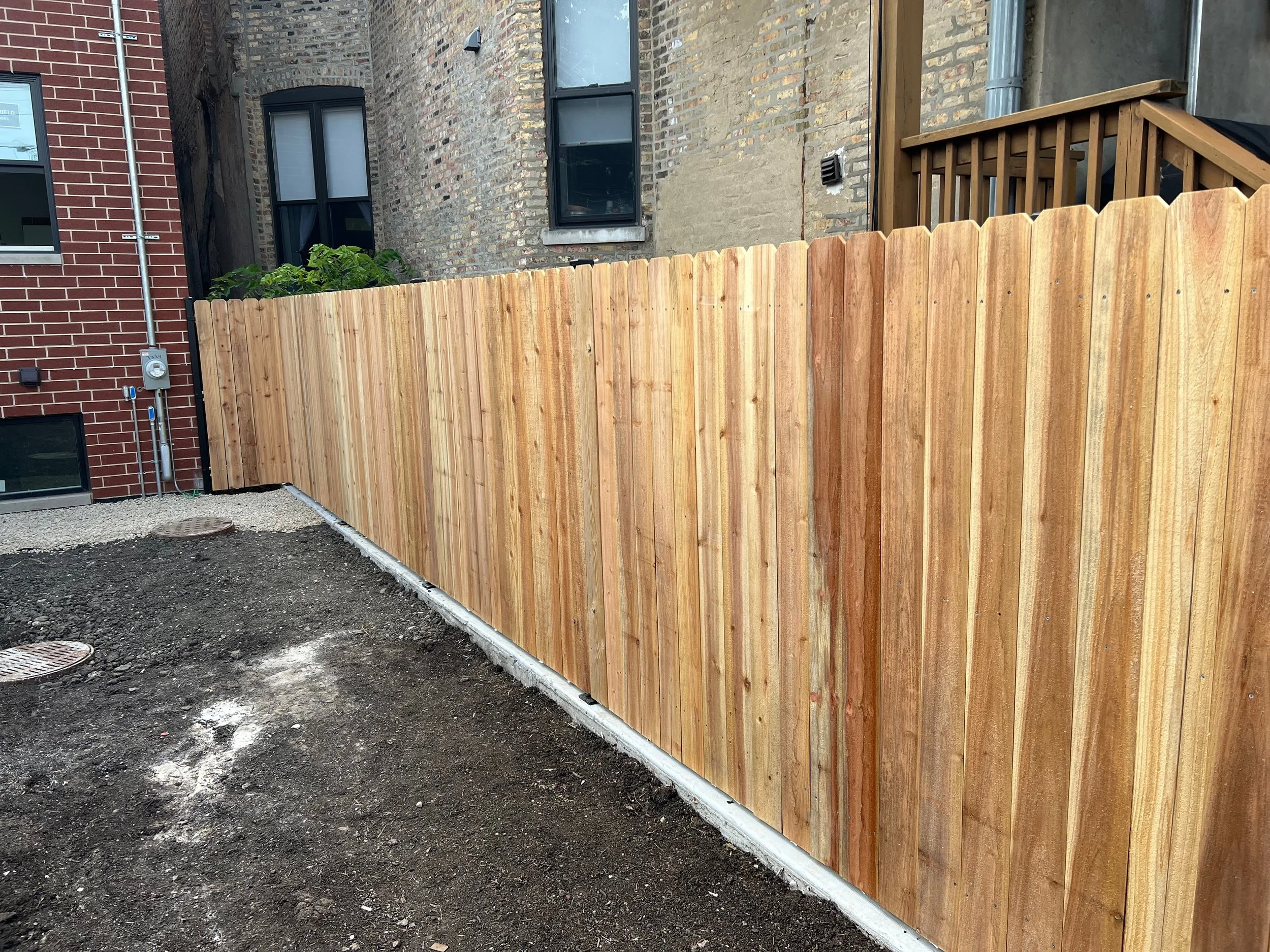 A backyard accessible with a tall wooden privacy fence, brick buildings with windows, an exposed brick wall, and an electrical meter with conduit on the side of a building. The ground is gravel and dirt.