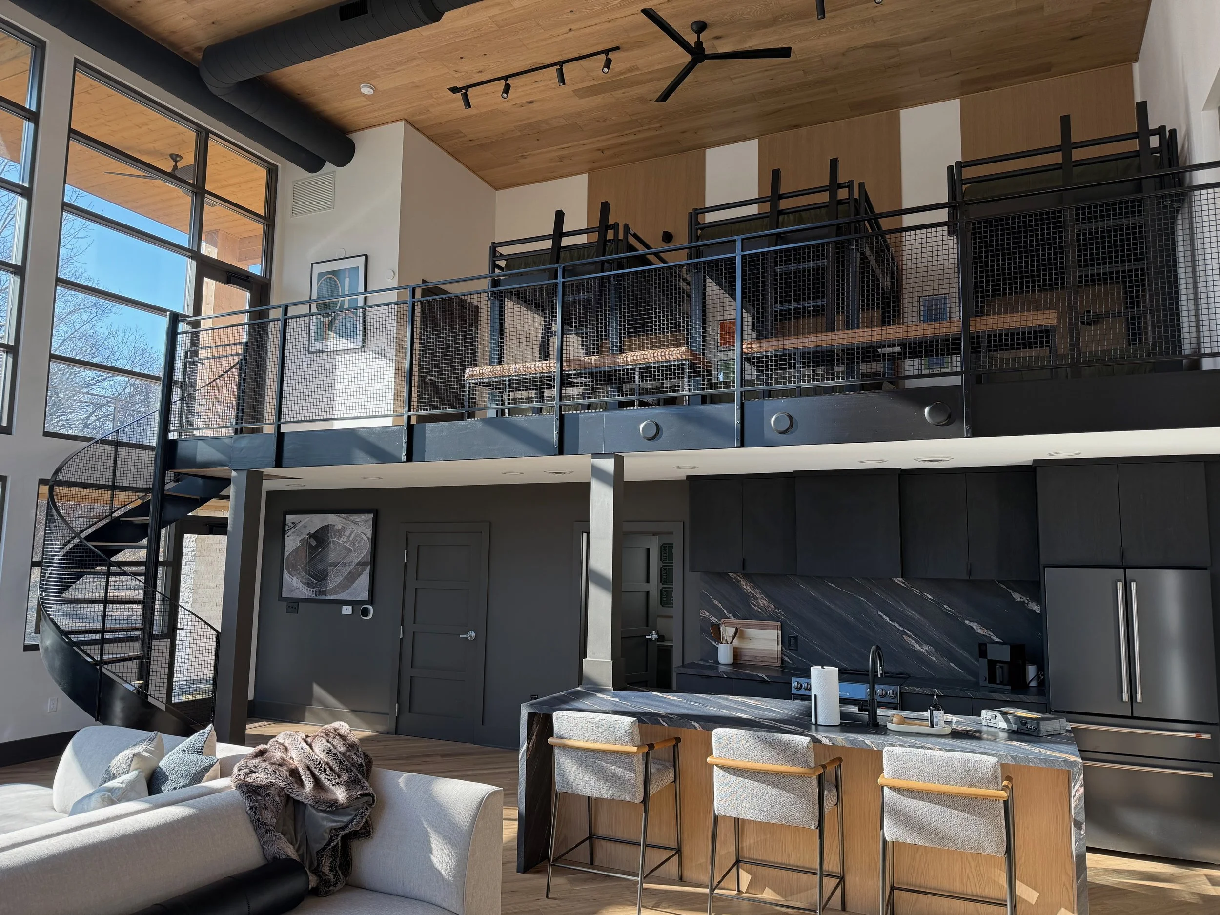 Open-concept living space with a kitchen, dining area, and upper-level balcony. Large windows letting in natural light, modern black railing, wooden accents, high ceiling with track lighting, spiral staircase, and contemporary furniture.