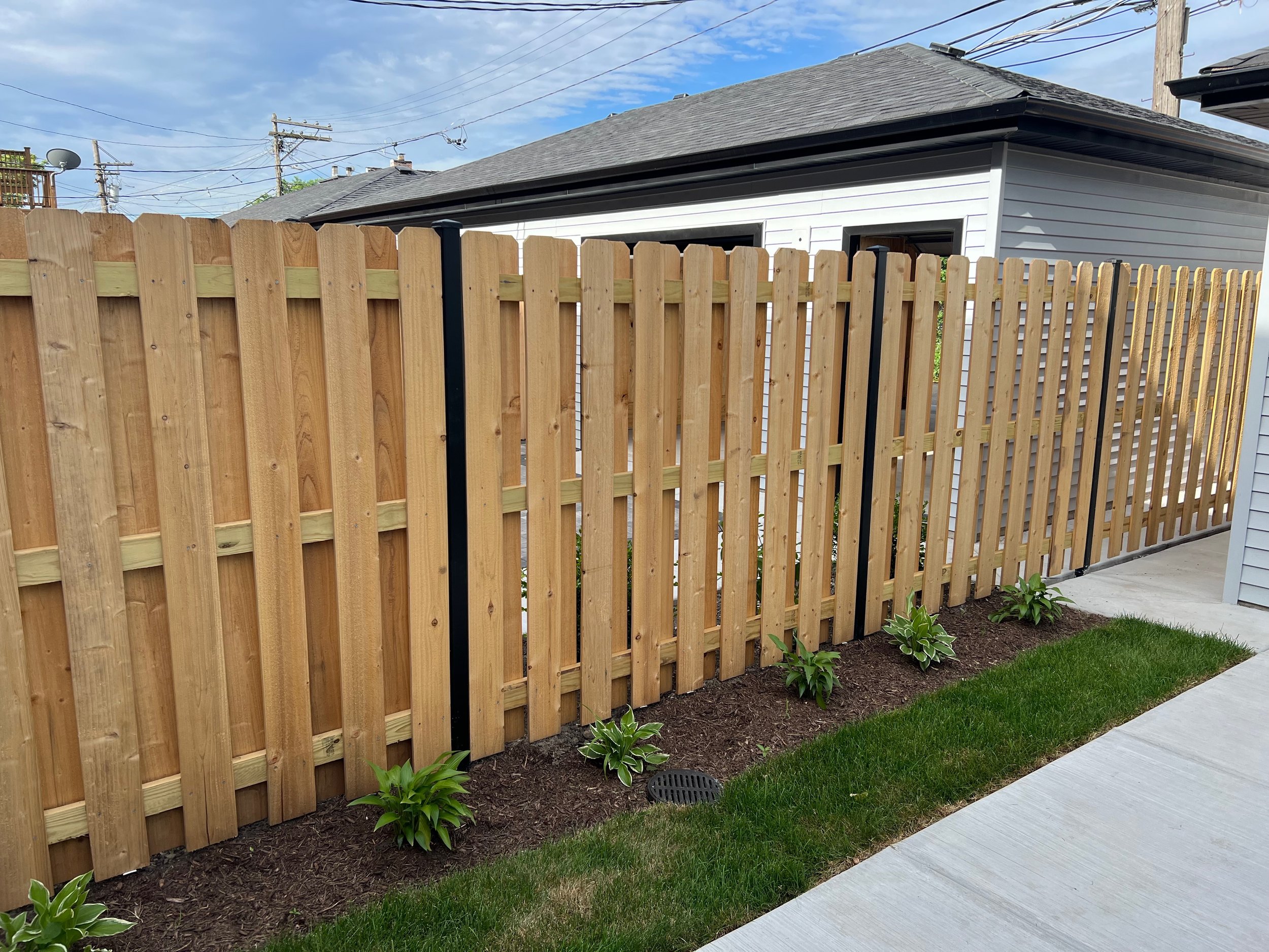 A new wooden fence borders a small garden with green plants, next to a concrete sidewalk and a white house with gray roof shingles.