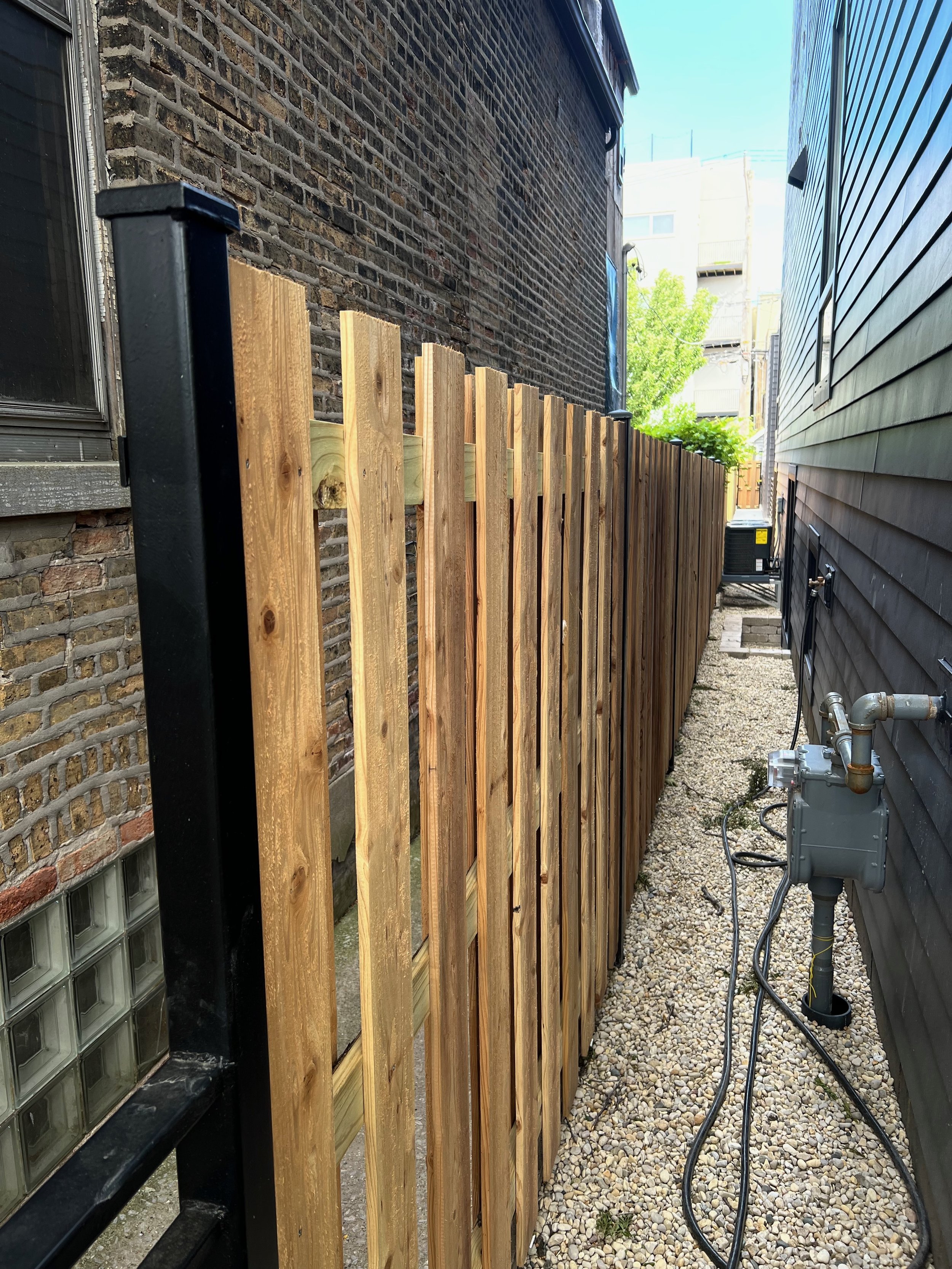 A narrow alleyway between two buildings with a newly installed wooden fence on the left and an existing black brick wall on the right. There are utility meters and gas pipes at the base of the right building, and an air conditioning unit further down