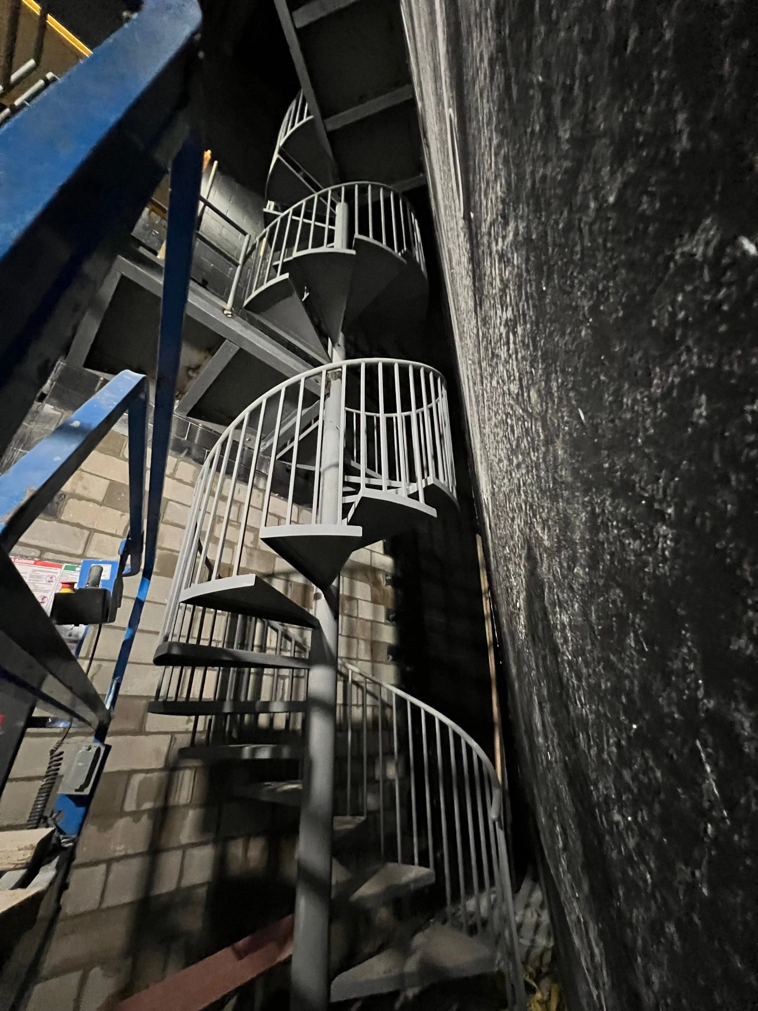 A spiral staircase with metal railings is located against a wall made of cinder blocks. The staircase is situated near a brick wall on the right side. The photo is taken at night or in a low-light setting.