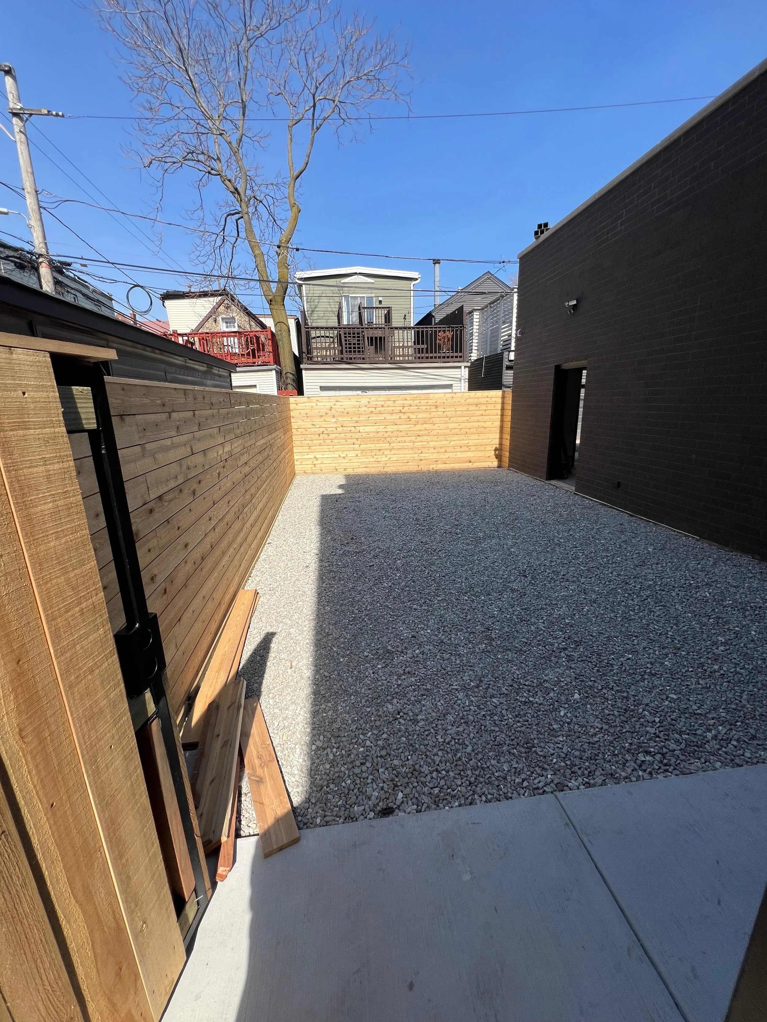 Residential backyard with a gravel surface, a tall wooden fence, and a black brick building wall on the right. Visible in the background are neighboring houses, a large leafless tree, power lines, and a clear blue sky.