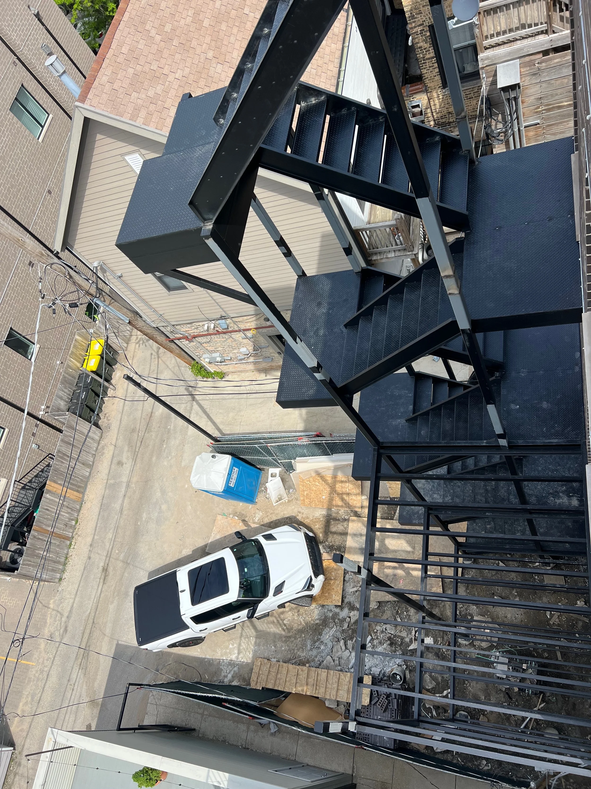 View of a narrow urban alleyway with parked white vehicle, a blue portable toilet, and a residential building with a brick fire escape staircase.